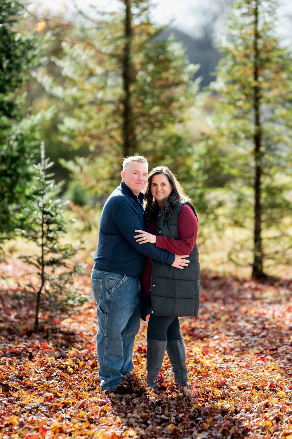 Happy couple embracing and smiling among pine trees with colorful fall foliage on the ground.