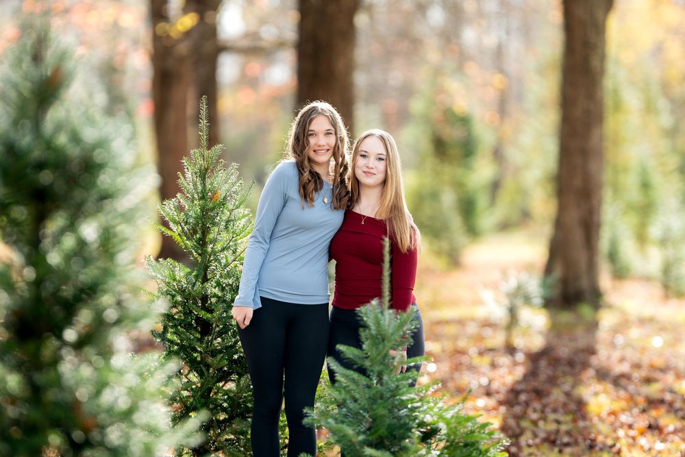 Two teenage girls posing together among Christmas trees in a sunlit autumn tree farm setting.