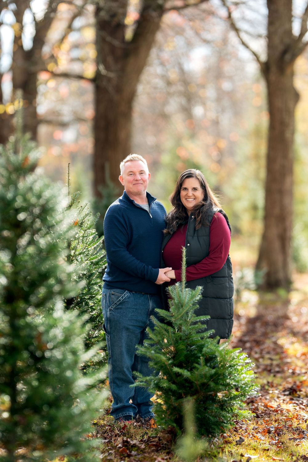 Smiling couple holding hands posing among young evergreen trees with autumn foliage backdrop.