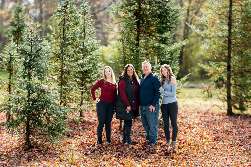 Family of four posing together among tall pine trees on a bed of autumn leaves.