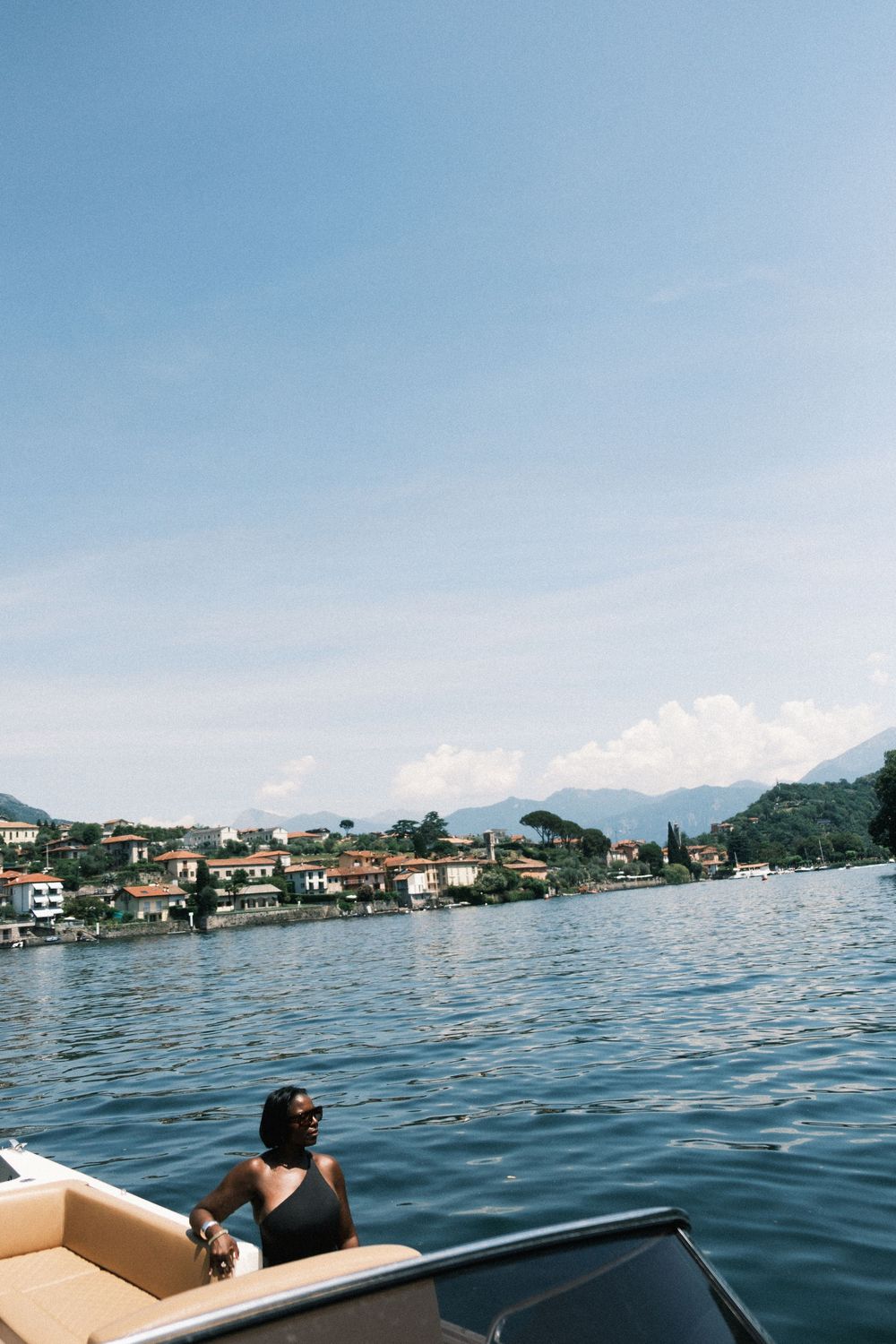 Woman in black swimsuit relaxing on a luxury boat with scenic Italian lake village and mountains in background.