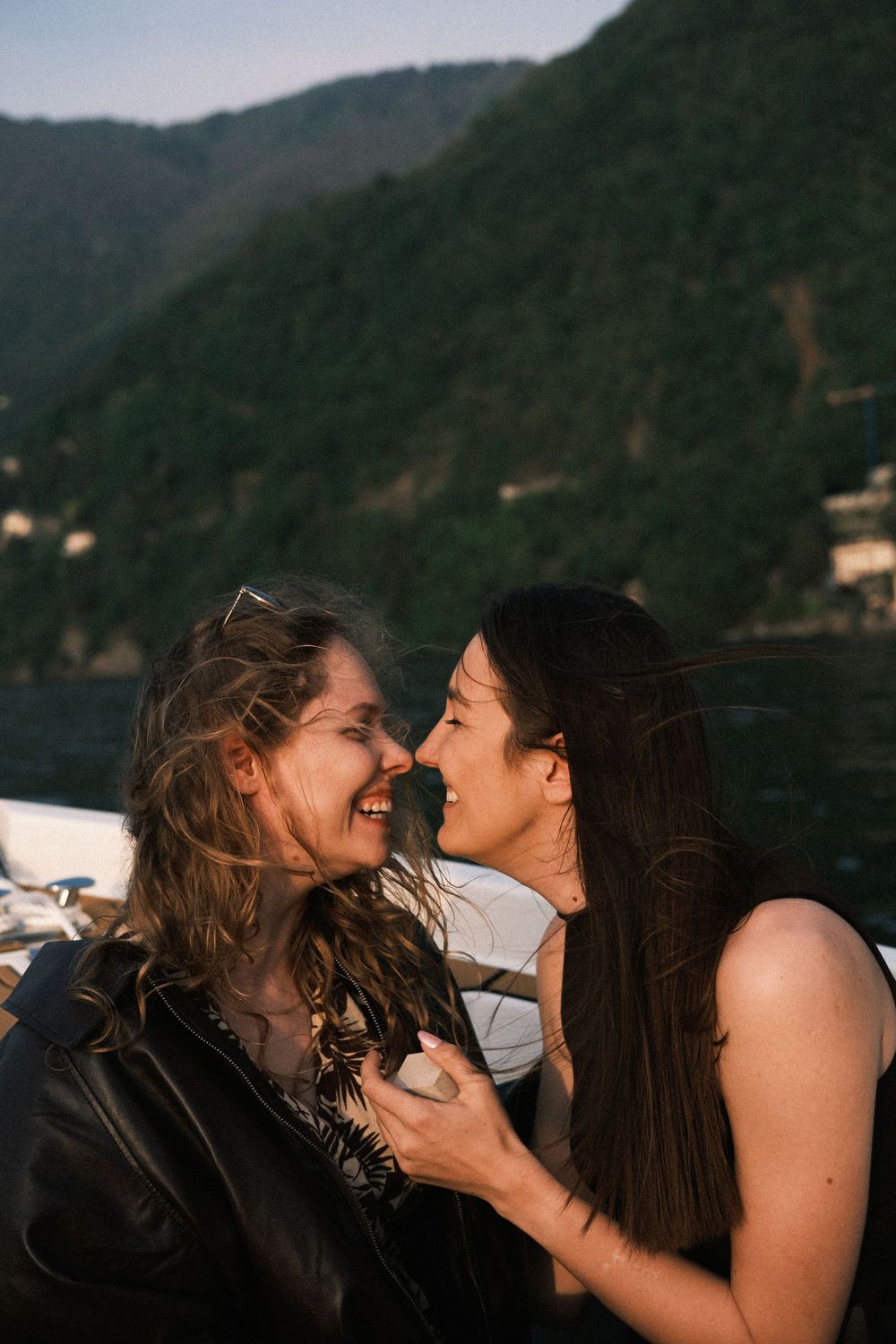 Two women share a joyful moment nose-to-nose on a boat with scenic green mountains and lake in background.