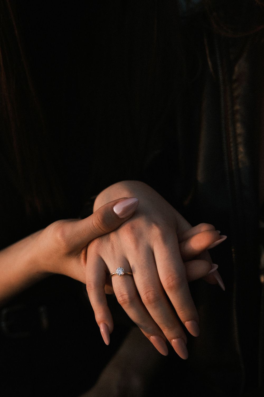 Close-up of elegant hands with nude nails wearing a sparkling diamond engagement ring against dark background.