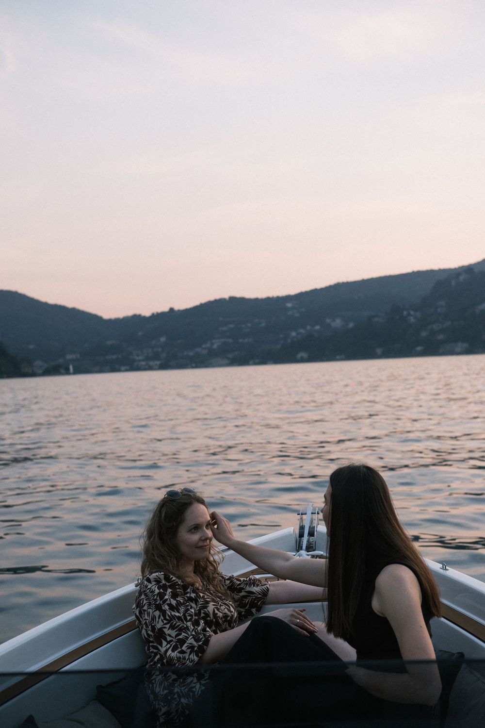 Two women sitting together in a rowboat on a calm lake at dusk with misty hills in the background.