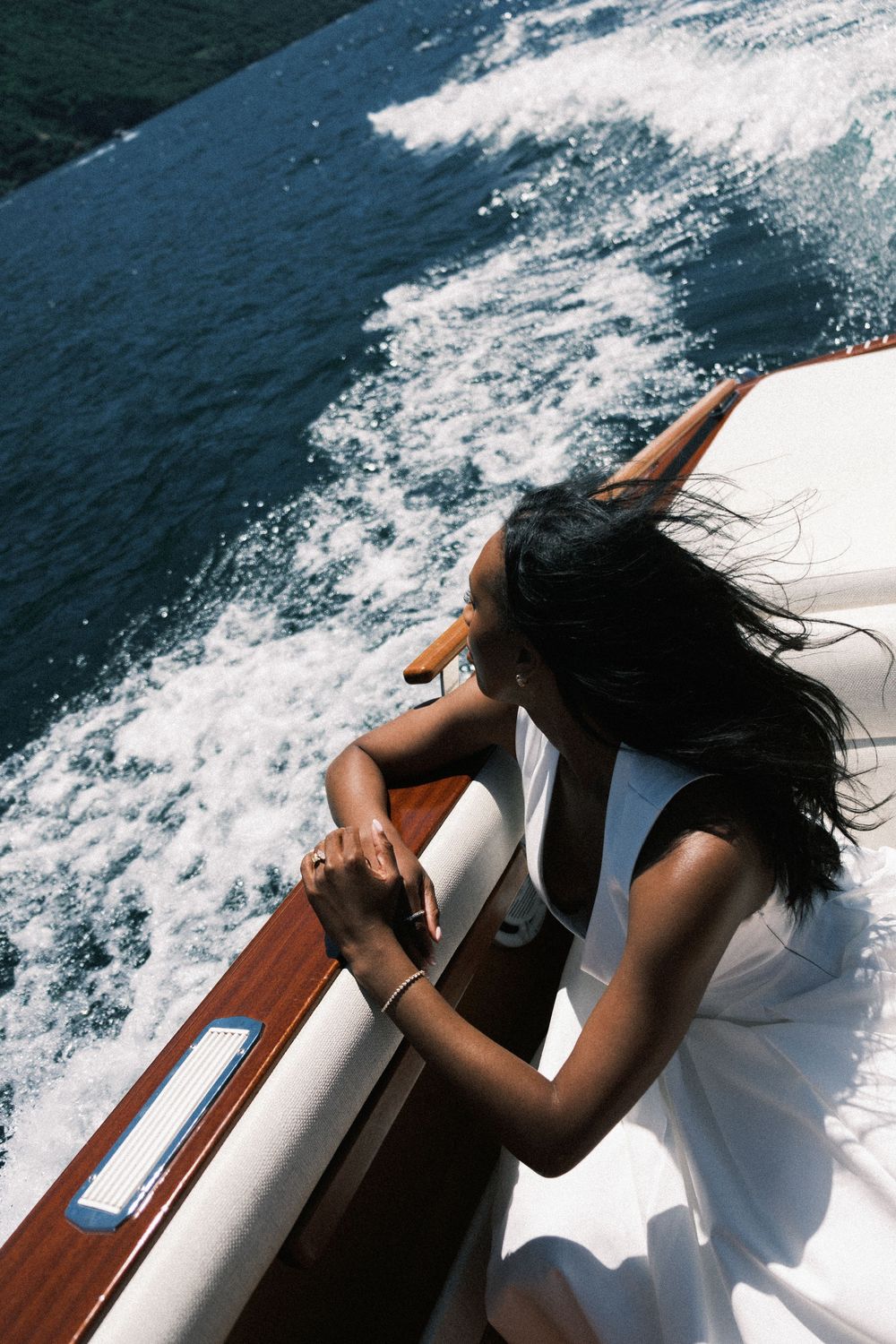 A woman with dark hair leans over a wooden boat railing, gazing at the ocean wake on a sunny day.