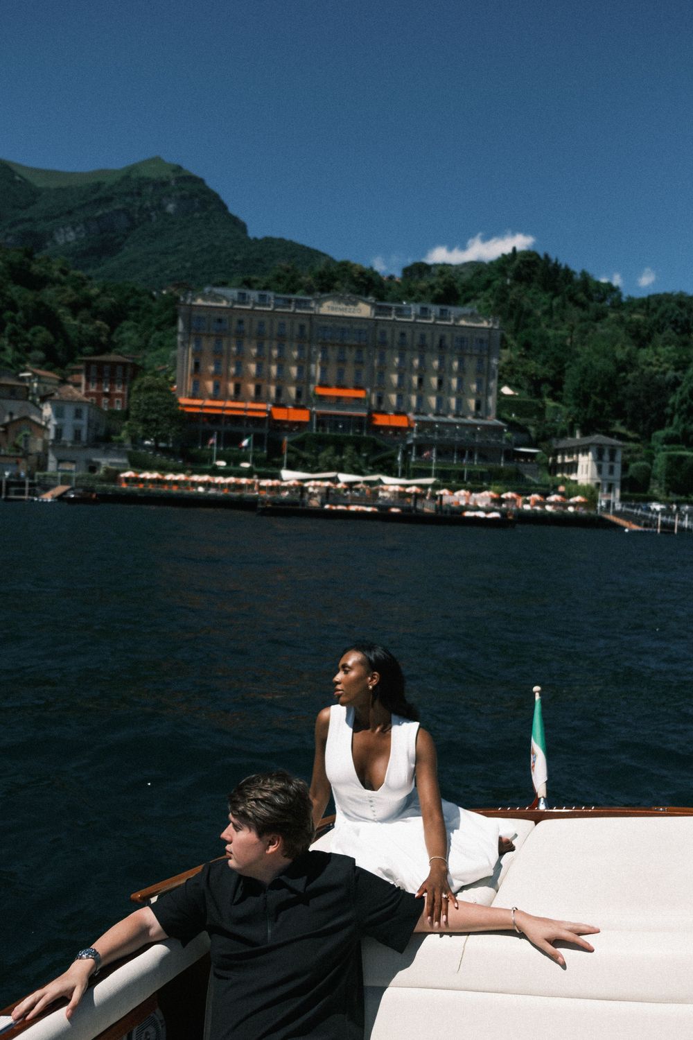 Two people on a wooden boat with a luxury lakeside hotel and green mountains in the background at Lake Como.