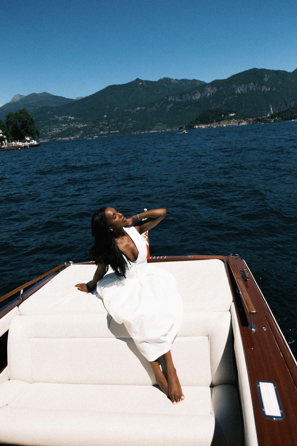 Woman in white dress relaxing on luxury boat bow with scenic mountain lake backdrop on sunny day.