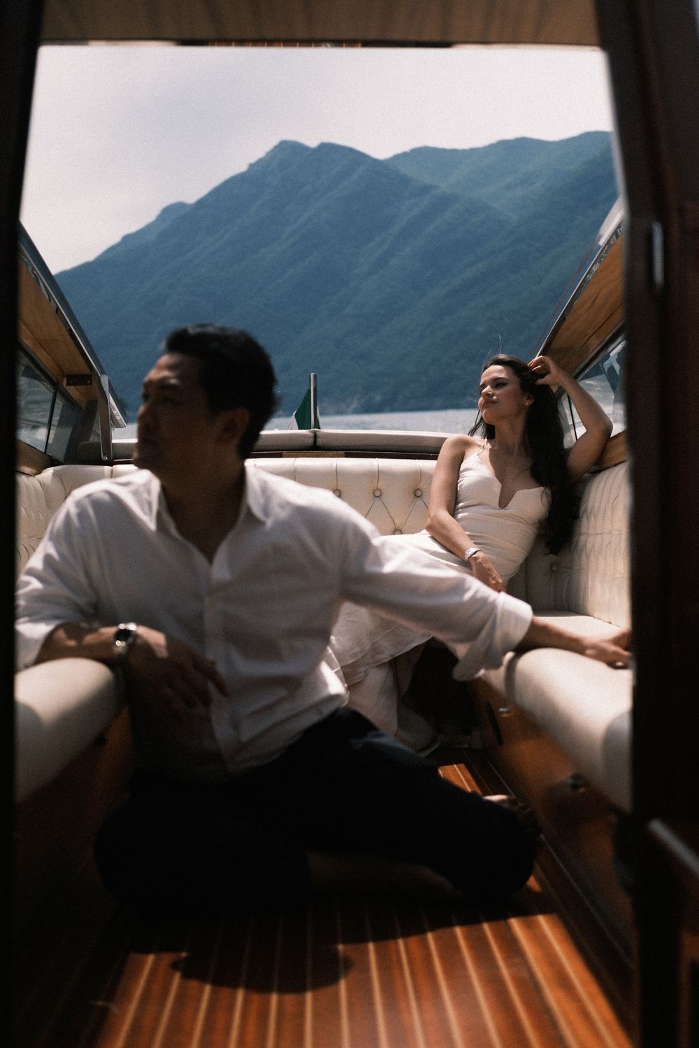 Couple relaxing on a luxury wooden boat with mountain lake views, man in white shirt seated on deck floor.