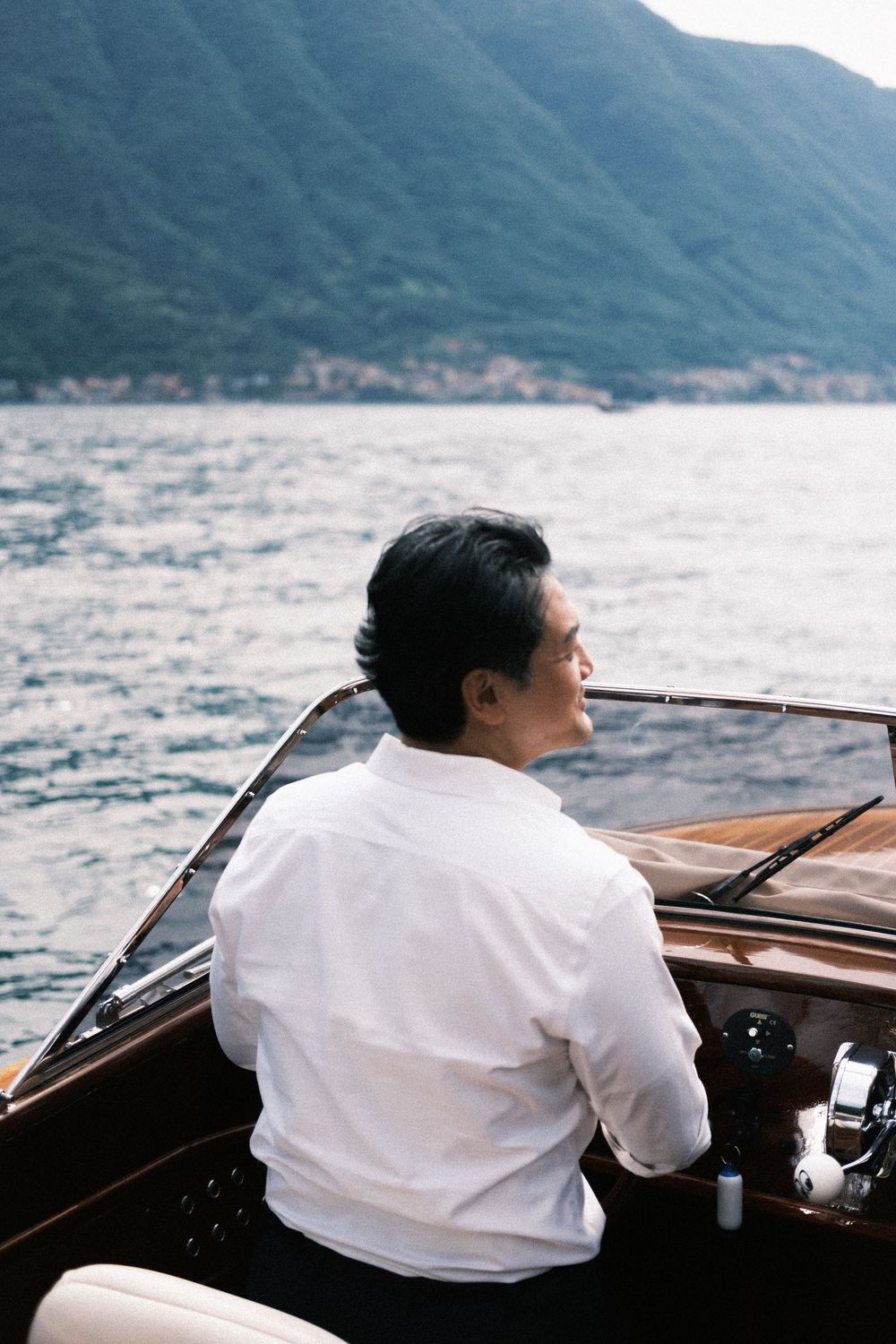 Man in white shirt sitting in a luxury wooden speedboat on a calm lake with mountain backdrop.