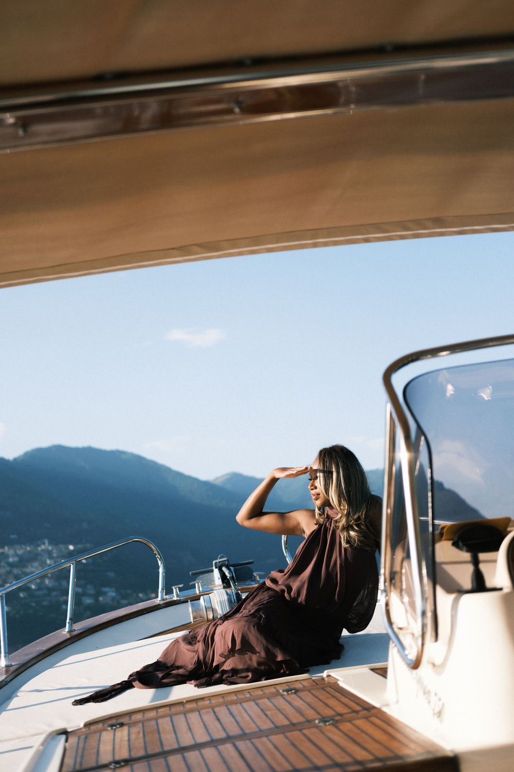 Woman in elegant dark dress sitting on yacht deck, shielding eyes from sun with mountain backdrop.