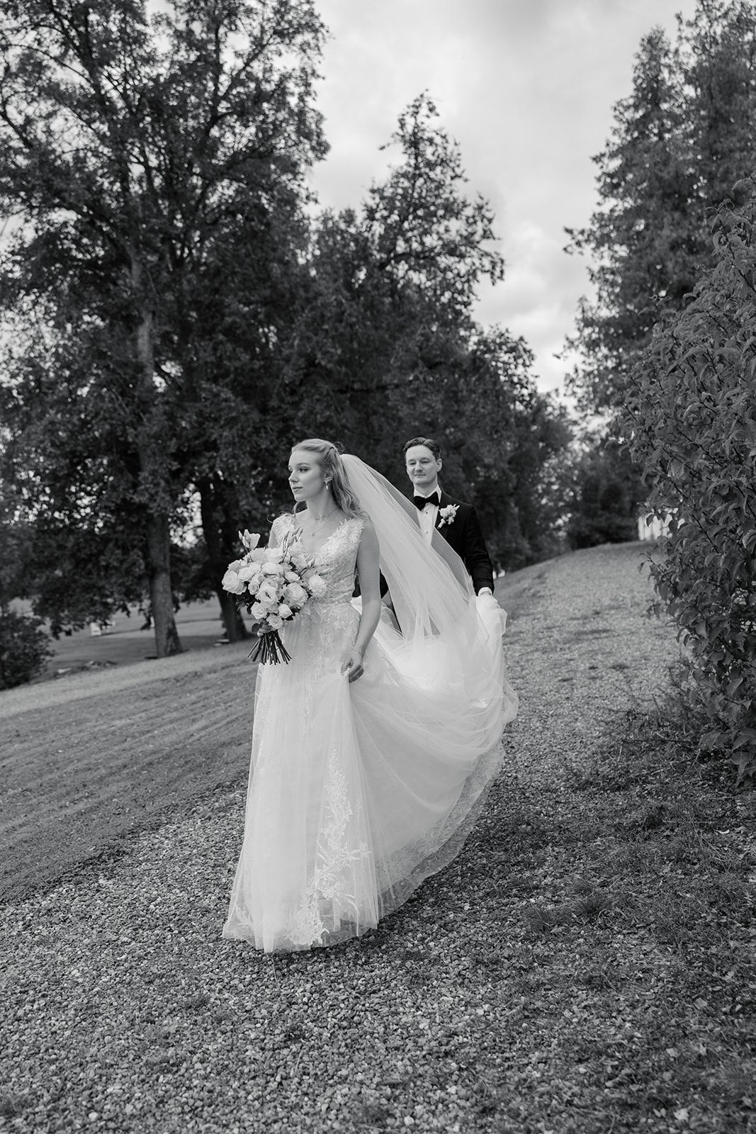 A couple in wedding attire walk together across a grassy hillside in black and white photography.