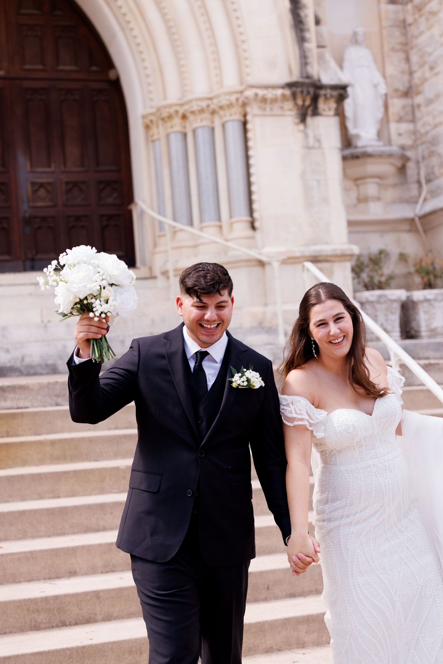 A joyful couple exits their wedding ceremony holding a white bouquet on church steps.