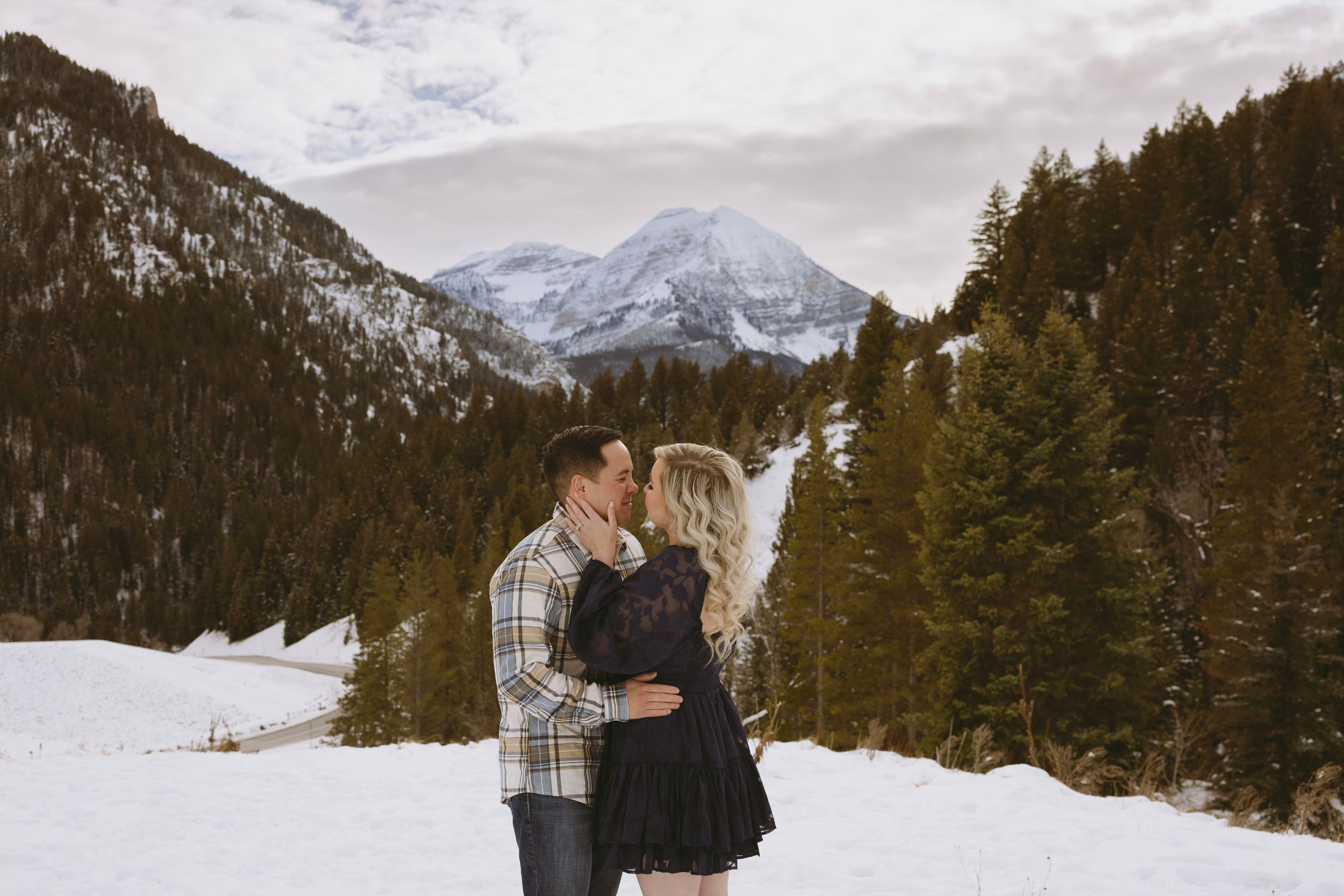 A couple embraces in a snowy winter mountain landscape with snow-covered evergreen trees and peaks in the background.