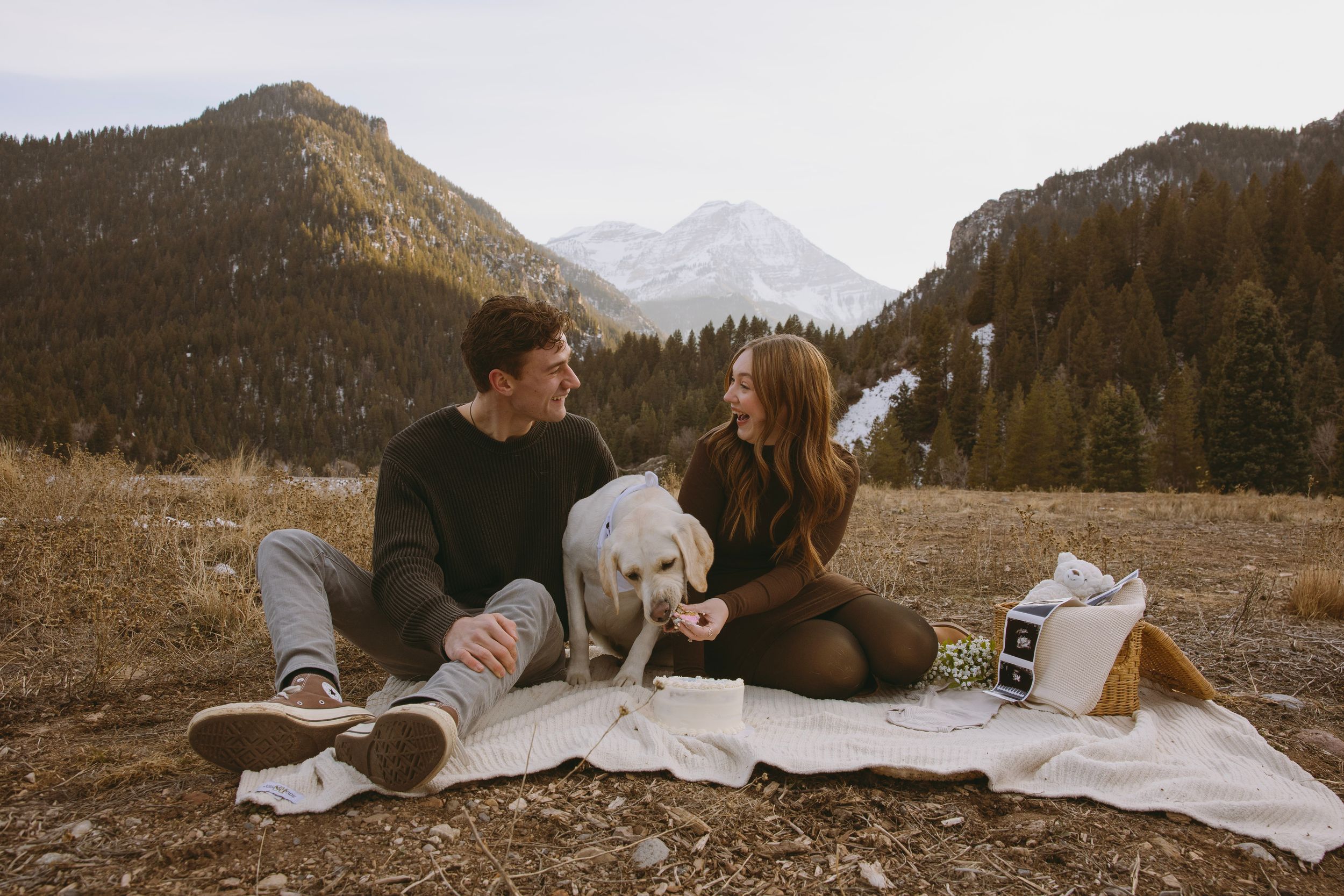 Couple sitting on blanket with white dog in mountain meadow, sharing a romantic picnic moment outdoors.