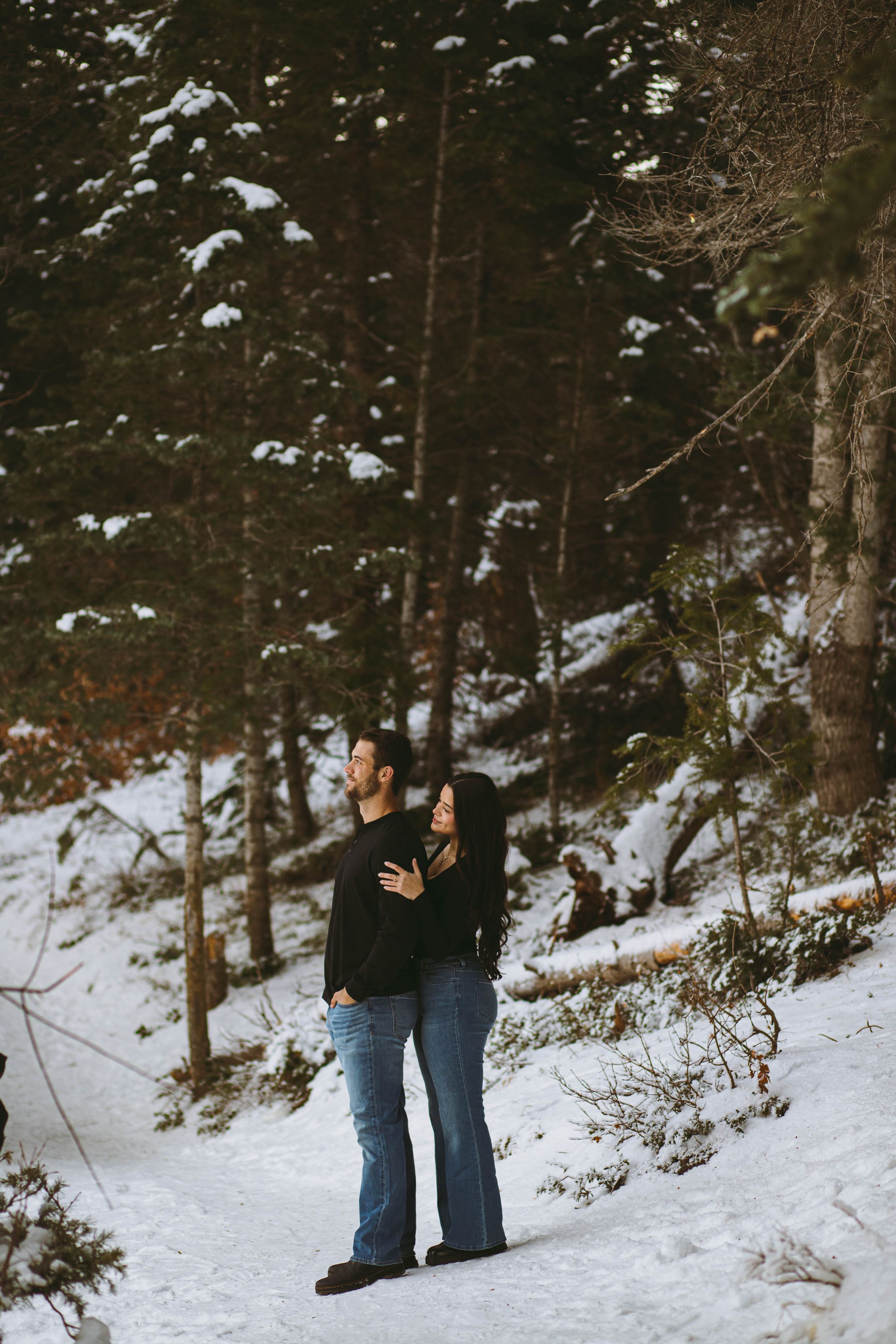 A couple embraces on a snowy forest path during winter, surrounded by snow-covered evergreen trees.
