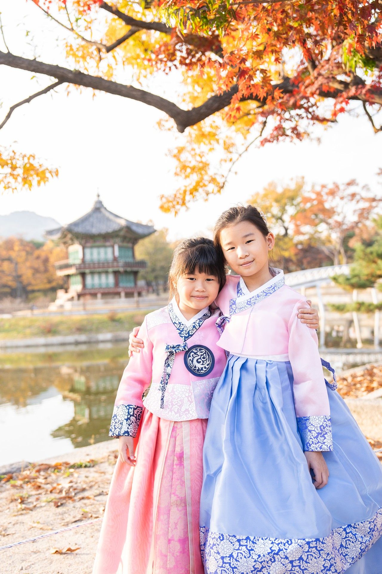 People wearing traditional Korean hanbok dresses pose among autumn foliage near a temple pavilion and lake.