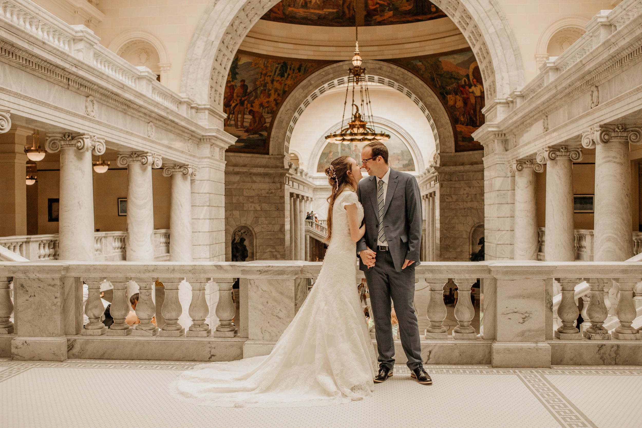 Wedding couple stands elegantly in Utah State Capitol rotunda with grand marble columns and architecture.