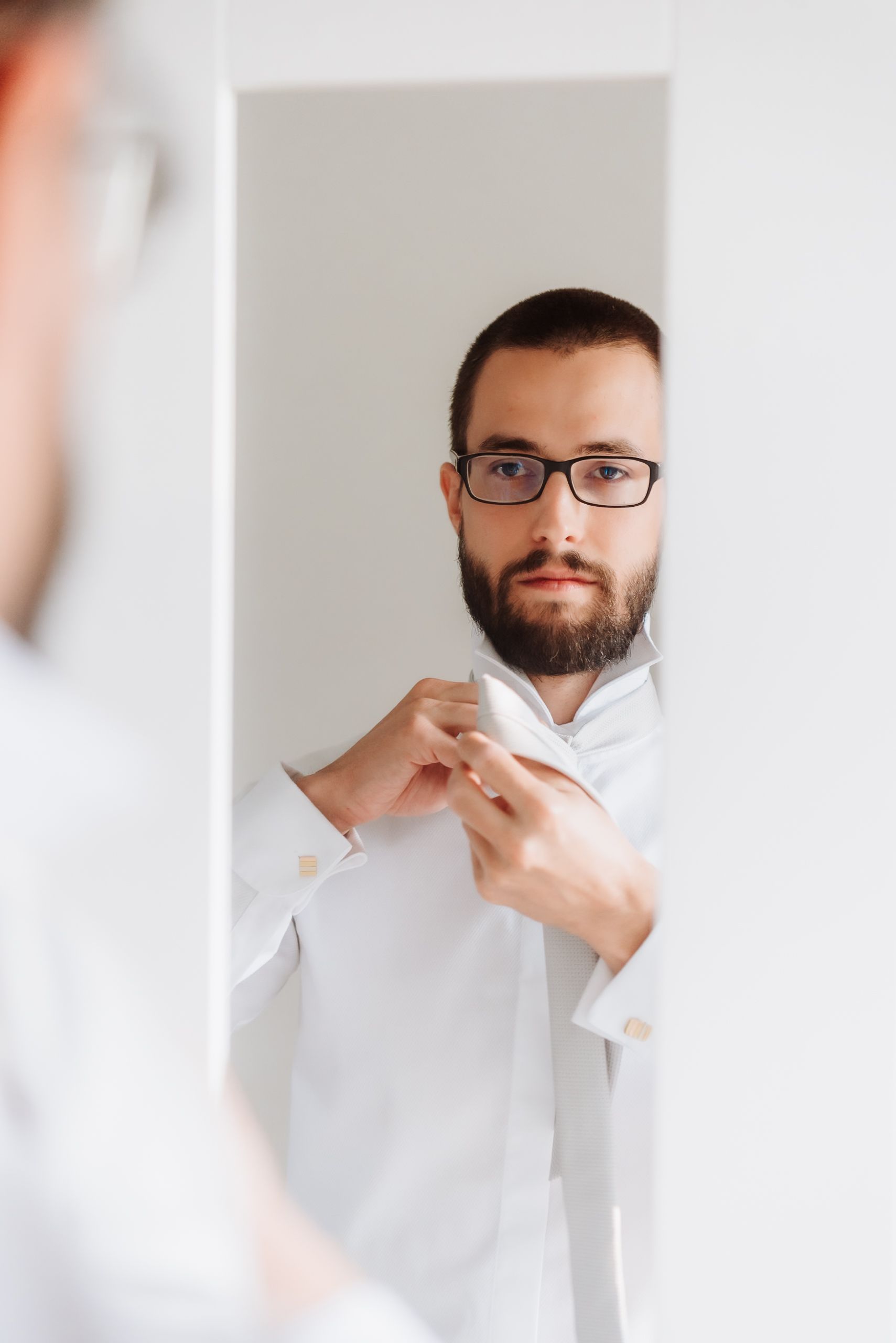 Person in white shirt and glasses adjusts their collar in a mirror reflection.
