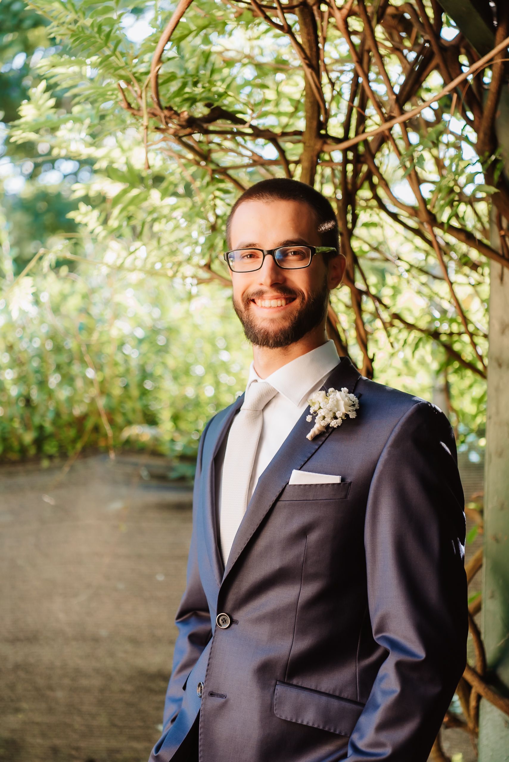A groom in a navy suit and glasses poses outdoors against a natural backdrop with greenery and trees.