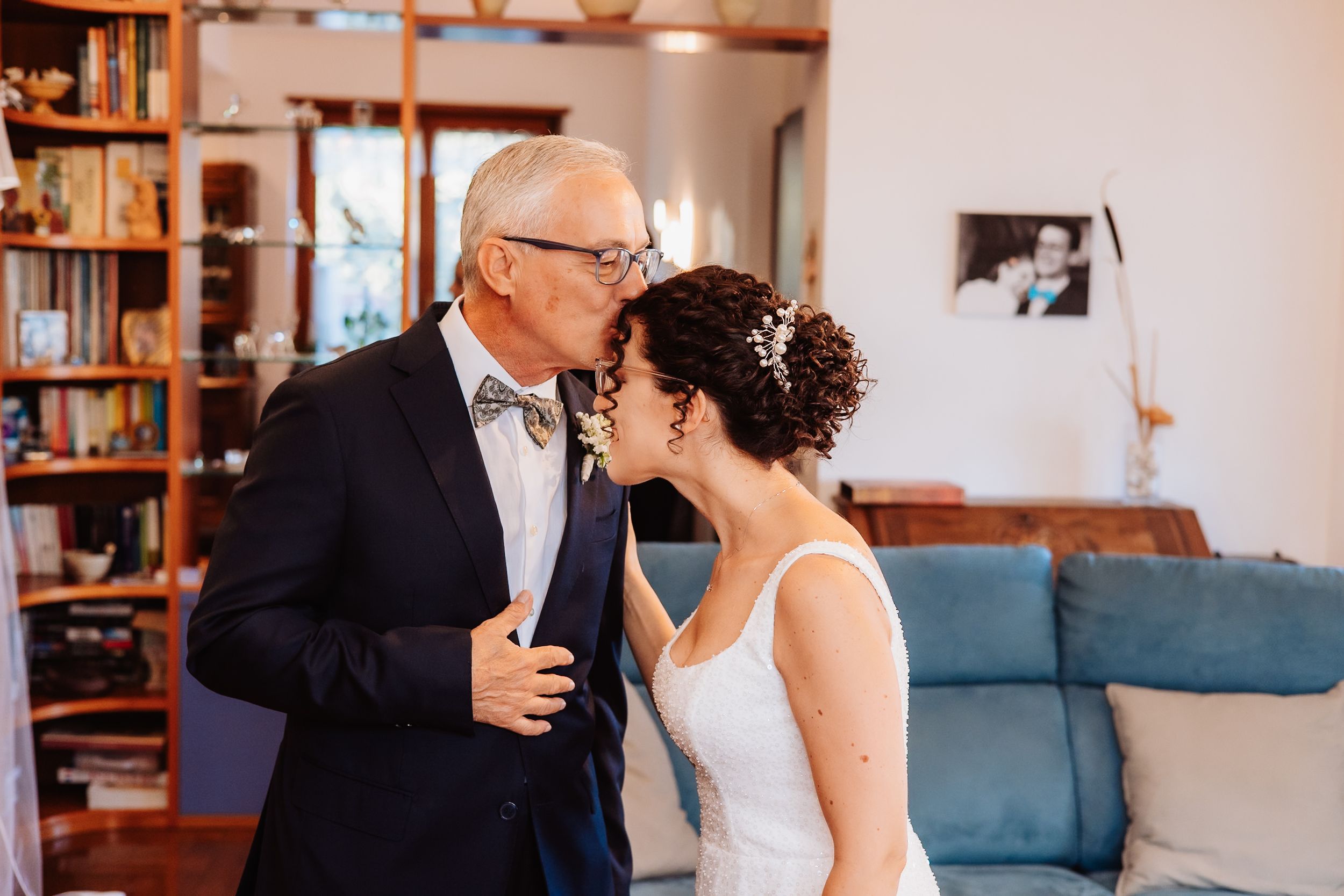 A couple shares a tender moment in a library setting with bookshelves and blue furniture visible in the background.