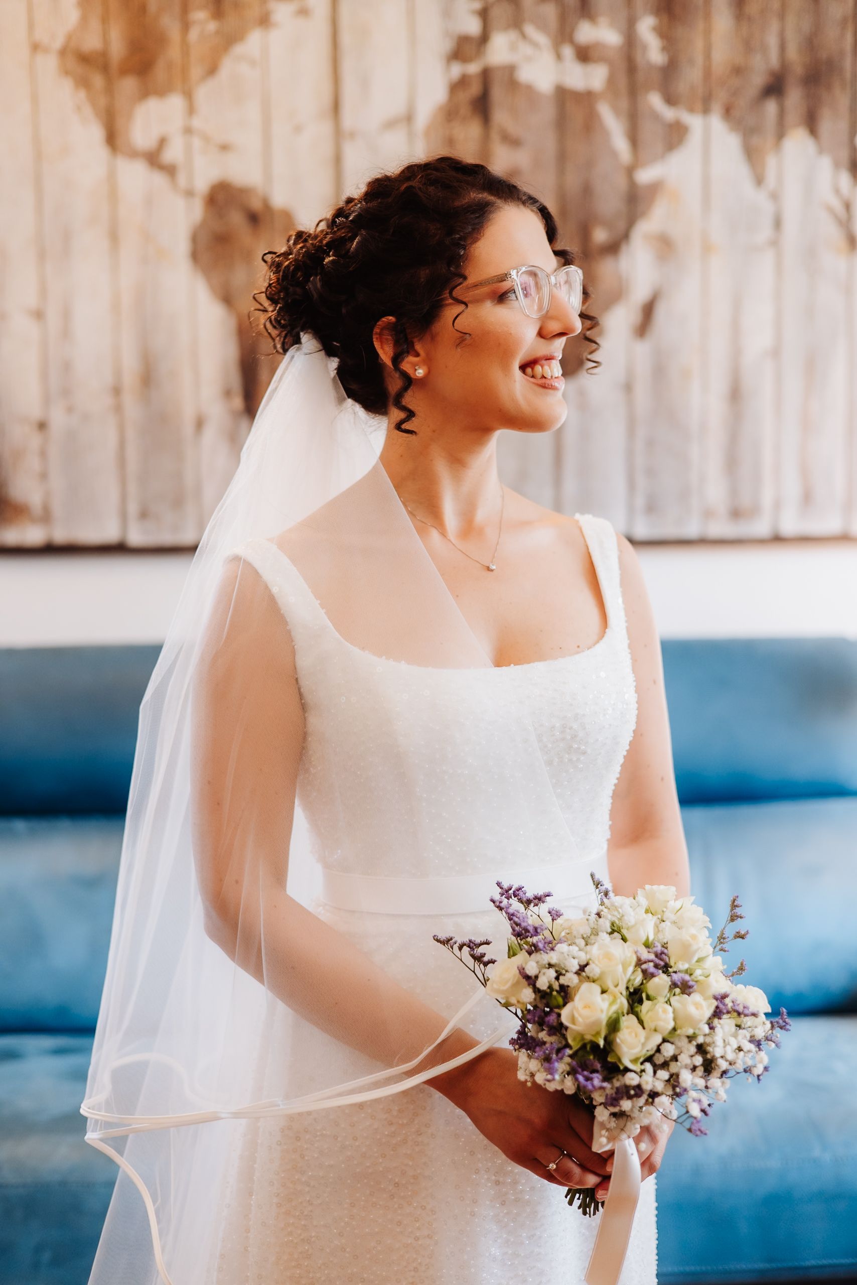A bride in a classic white wedding dress holds a delicate bouquet of white and purple flowers against a rustic backdrop.