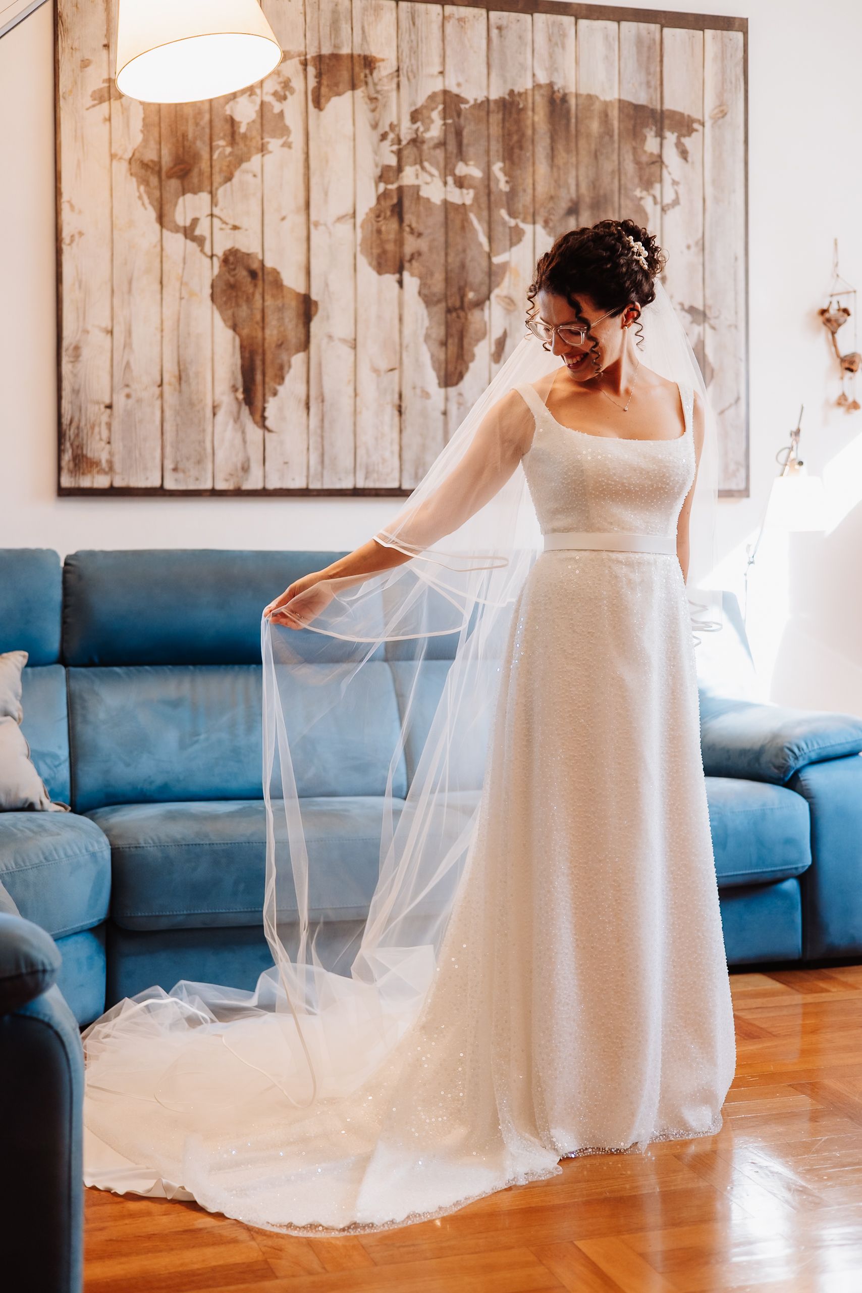 A flowing white wedding dress with a long veil is showcased in front of a wooden world map wall art and blue couch.