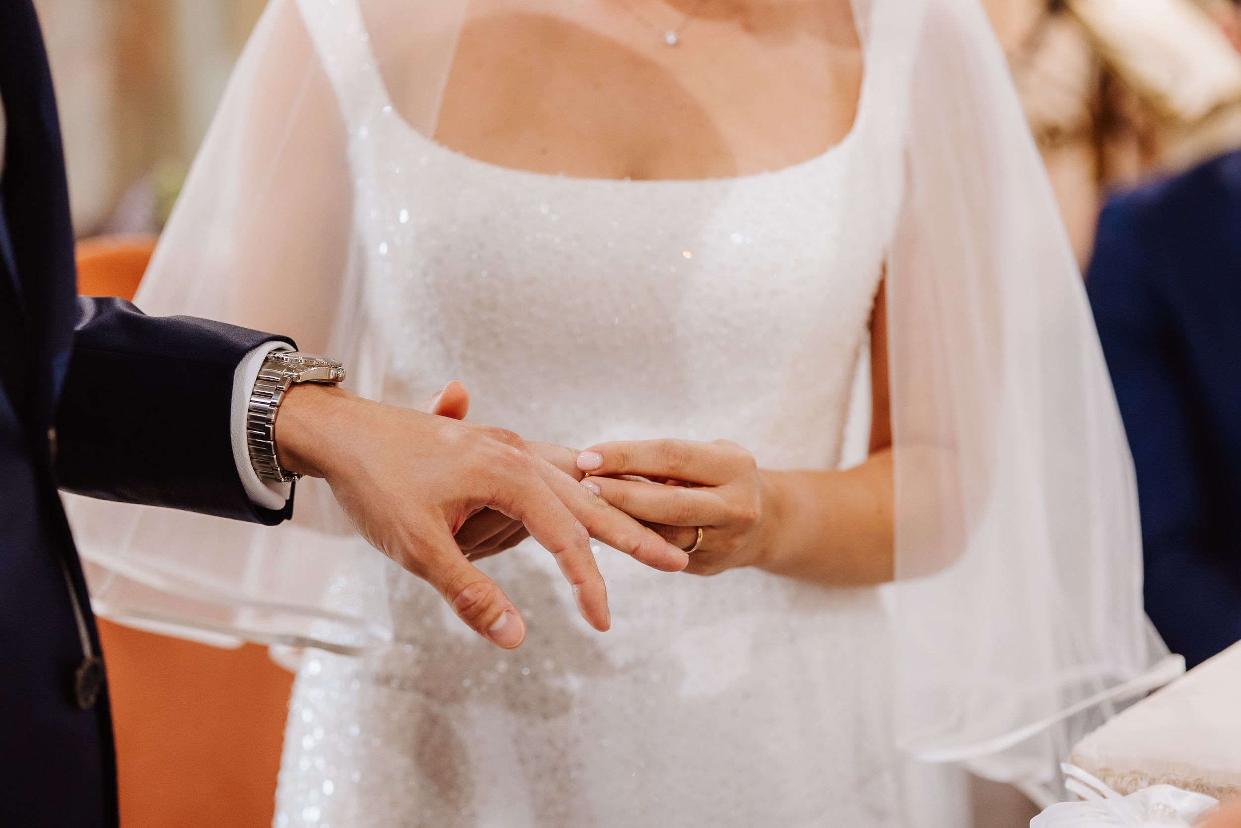 Close-up shot of hands exchanging wedding rings during a marriage ceremony with a white bridal dress in view.