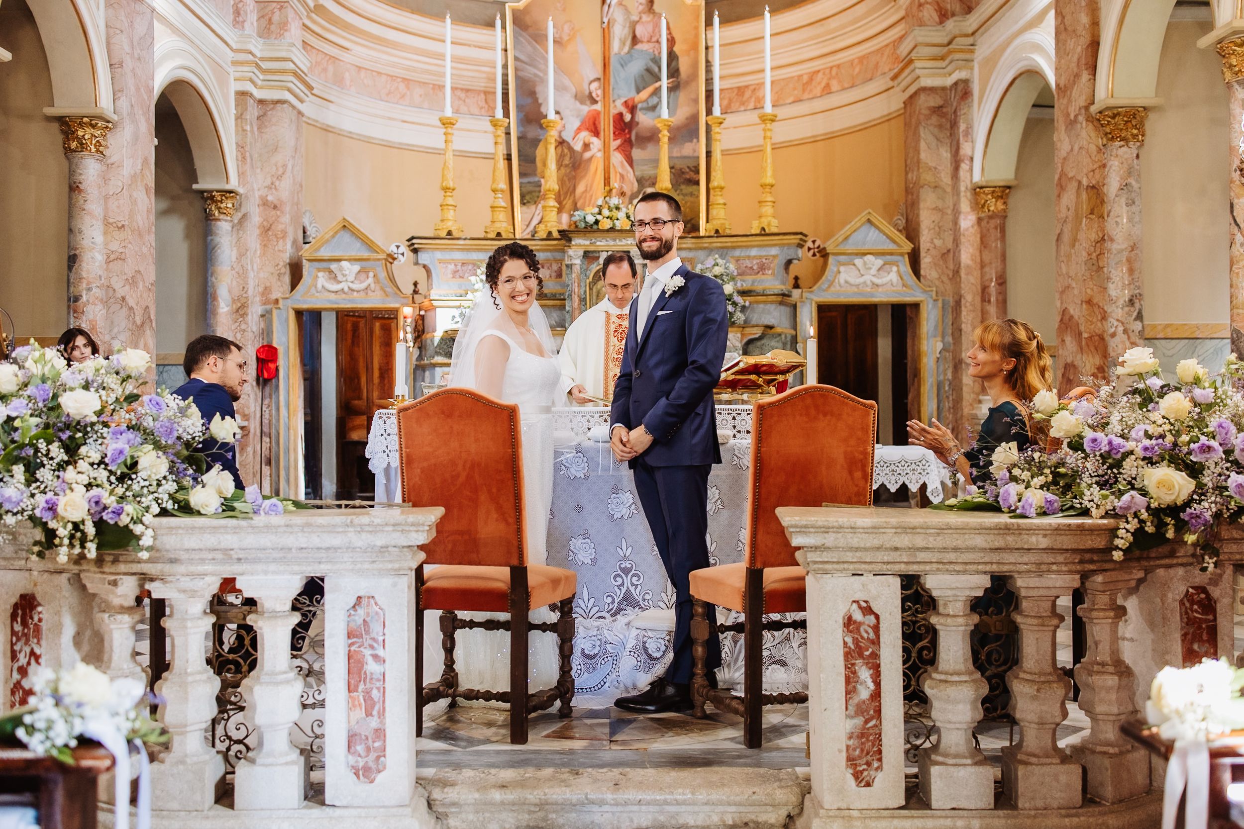 Wedding ceremony taking place in an elegant baroque chapel decorated with white floral arrangements and candles.