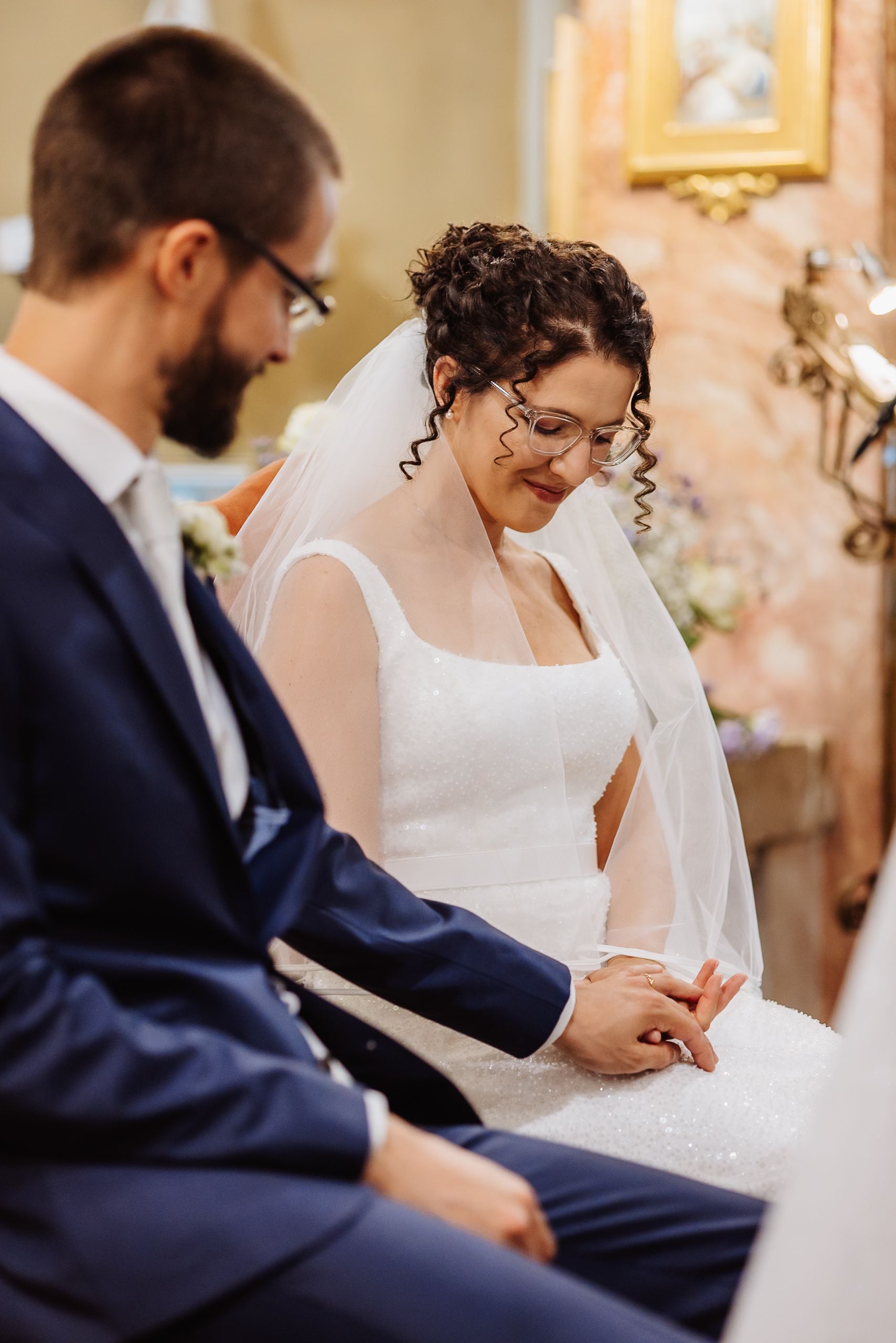 A couple sits together during a wedding ceremony, with the bride in white and groom in navy blue attire.