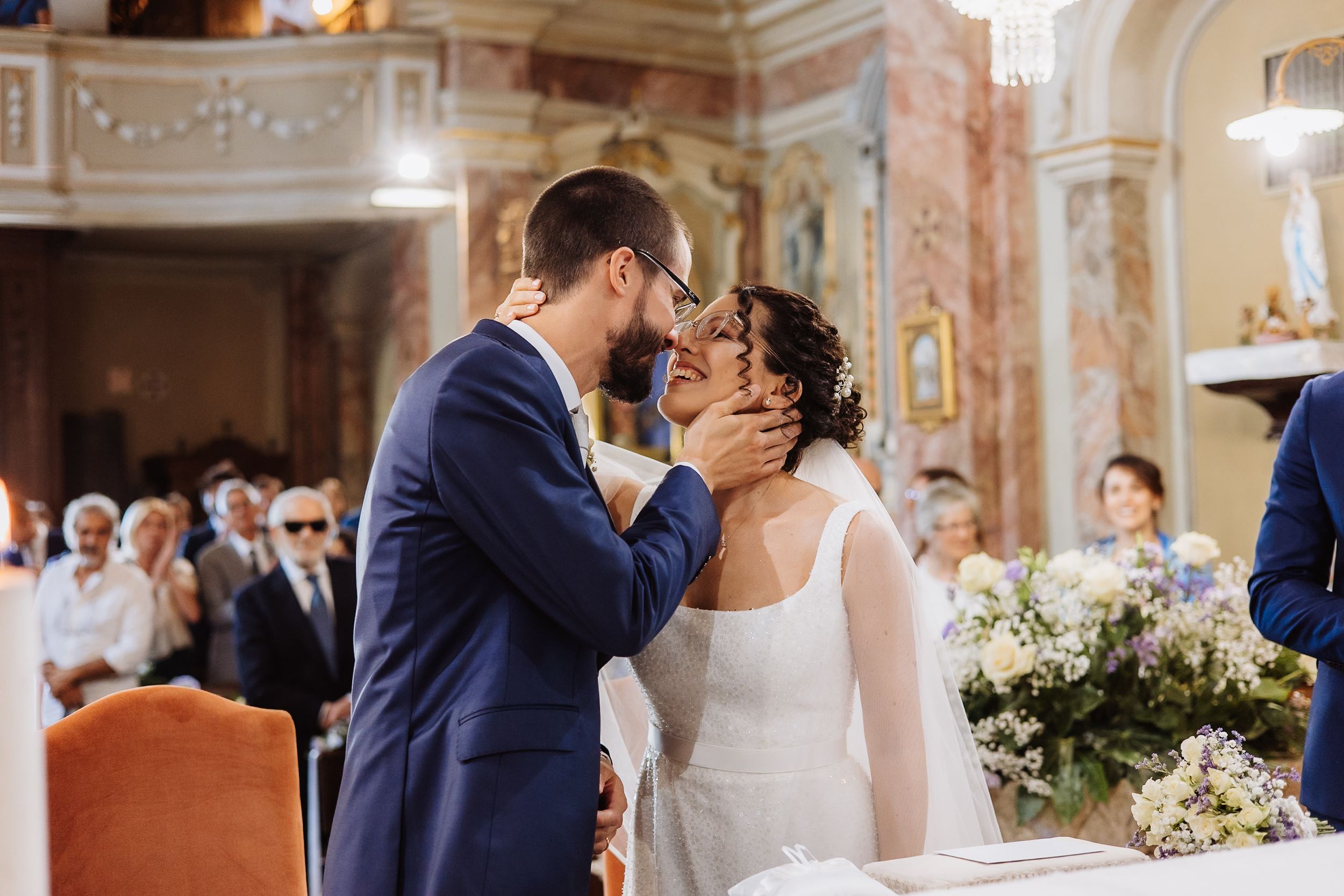 Couple shares romantic first kiss during elegant church wedding ceremony with ornate baroque interior.