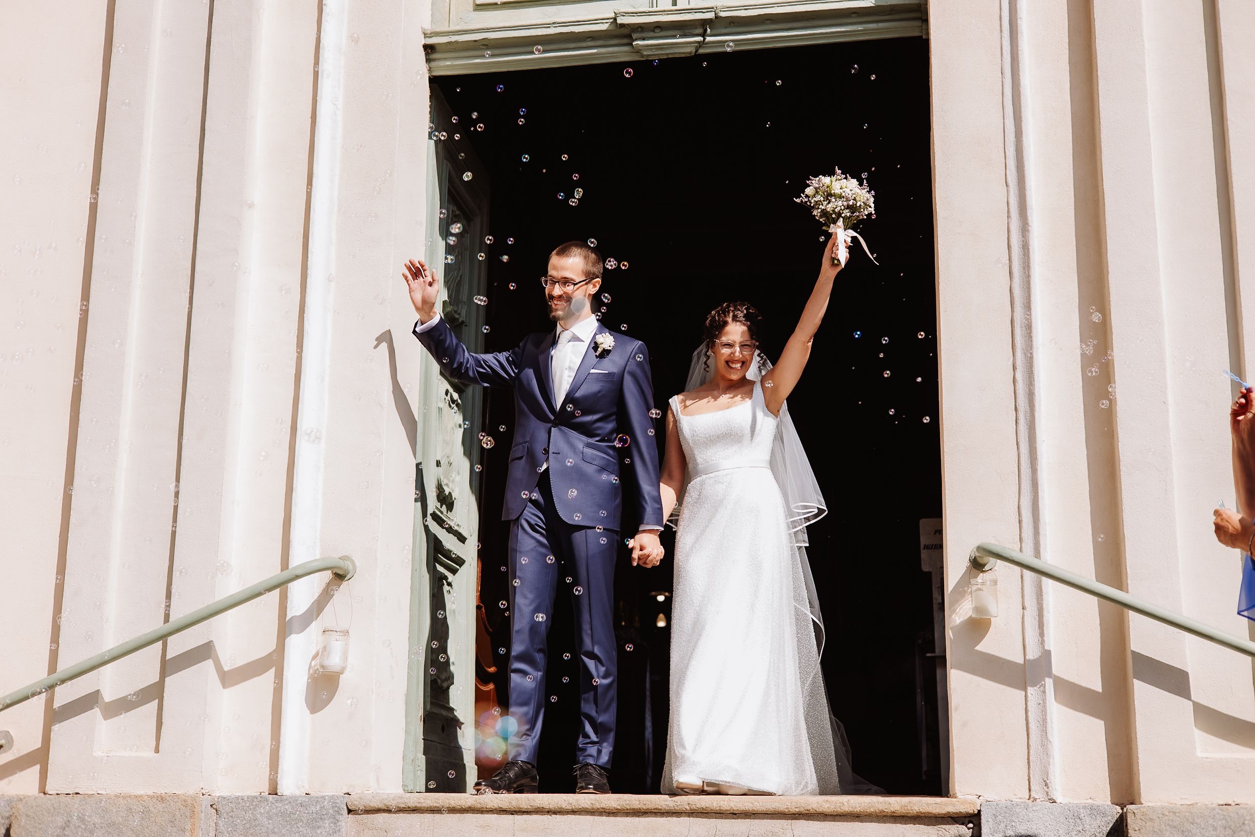 Wedding couple exits white church doors while guests throw rice in celebration on their special day.