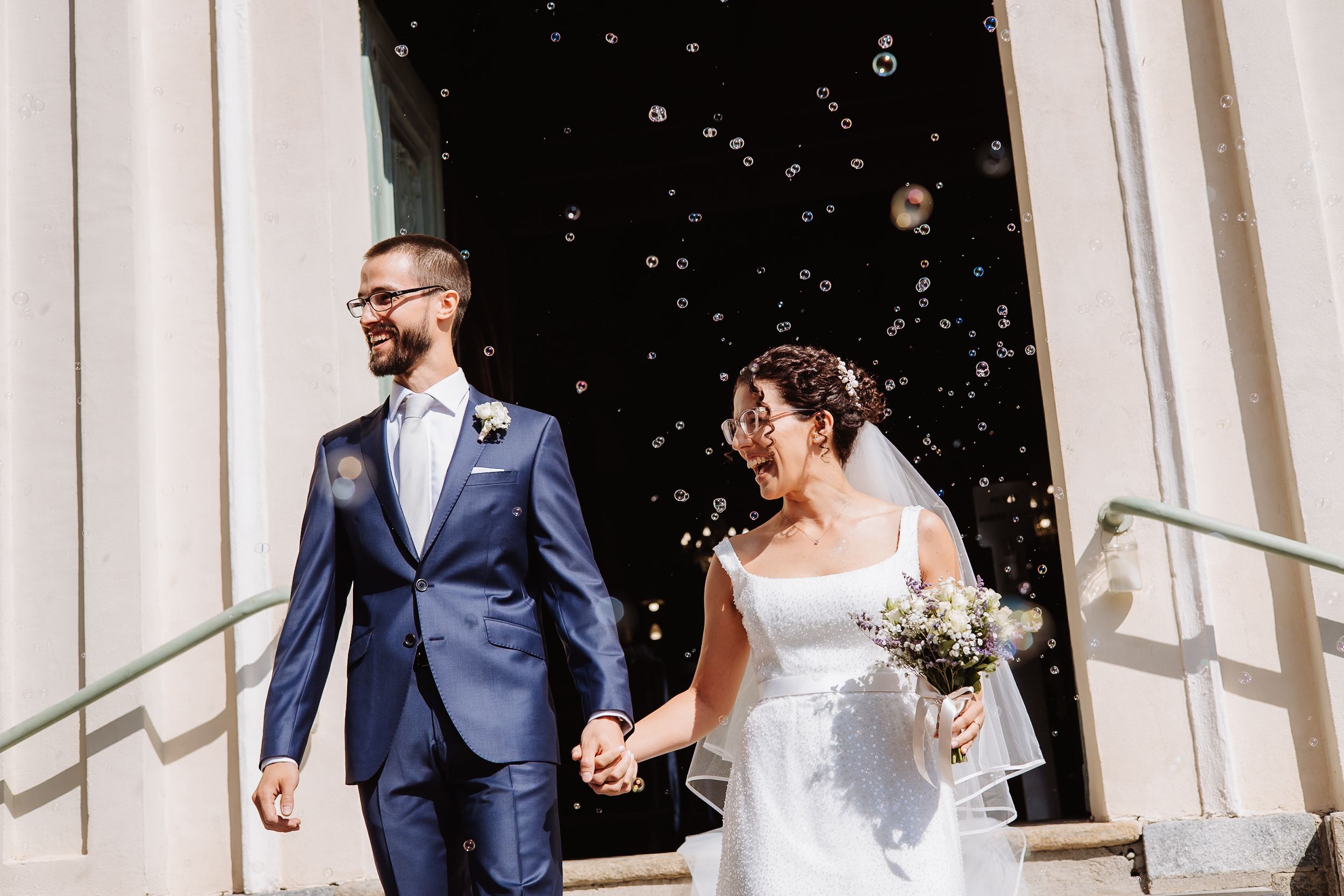 Wedding party walks and laughs together in front of white columns on a sunny day with confetti falling.