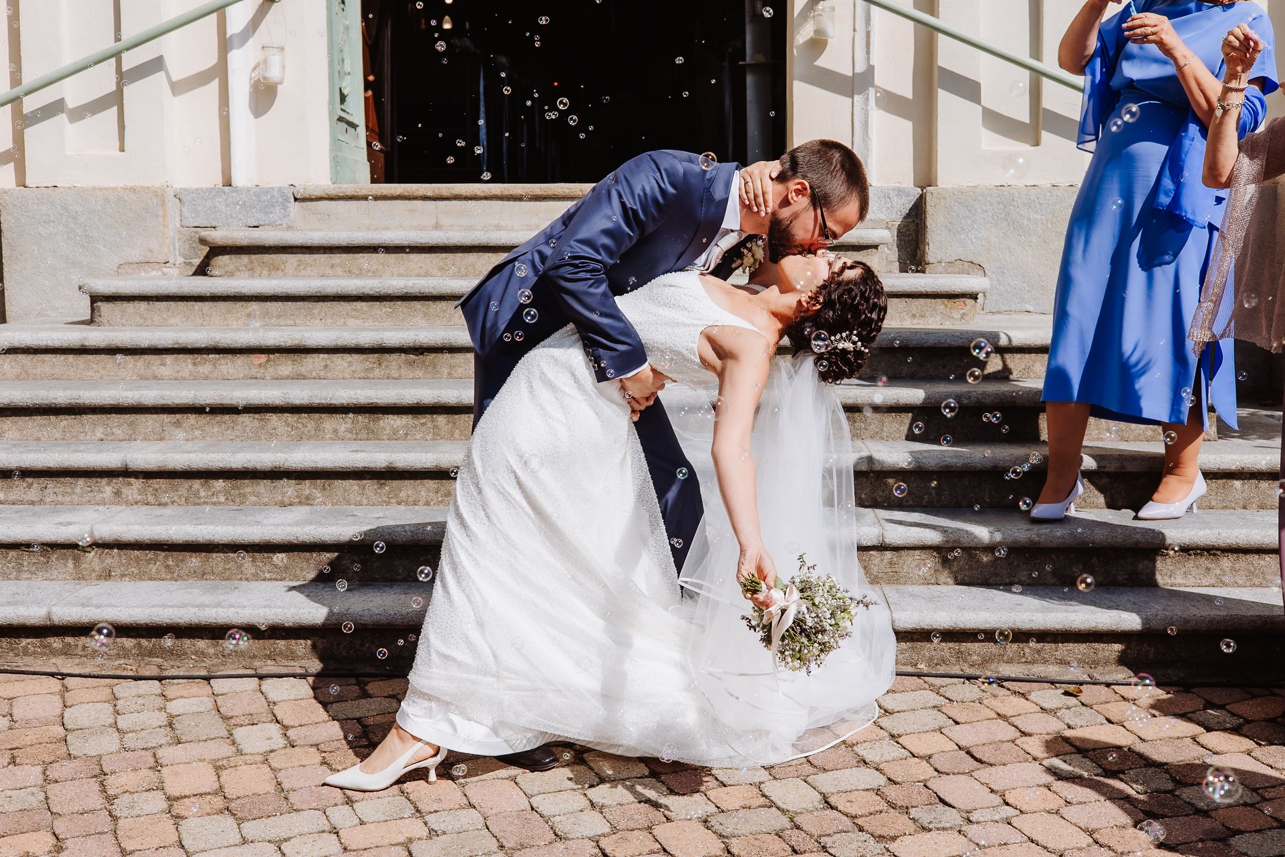A couple shares a romantic dip kiss on stone steps while someone in a blue dress looks on.