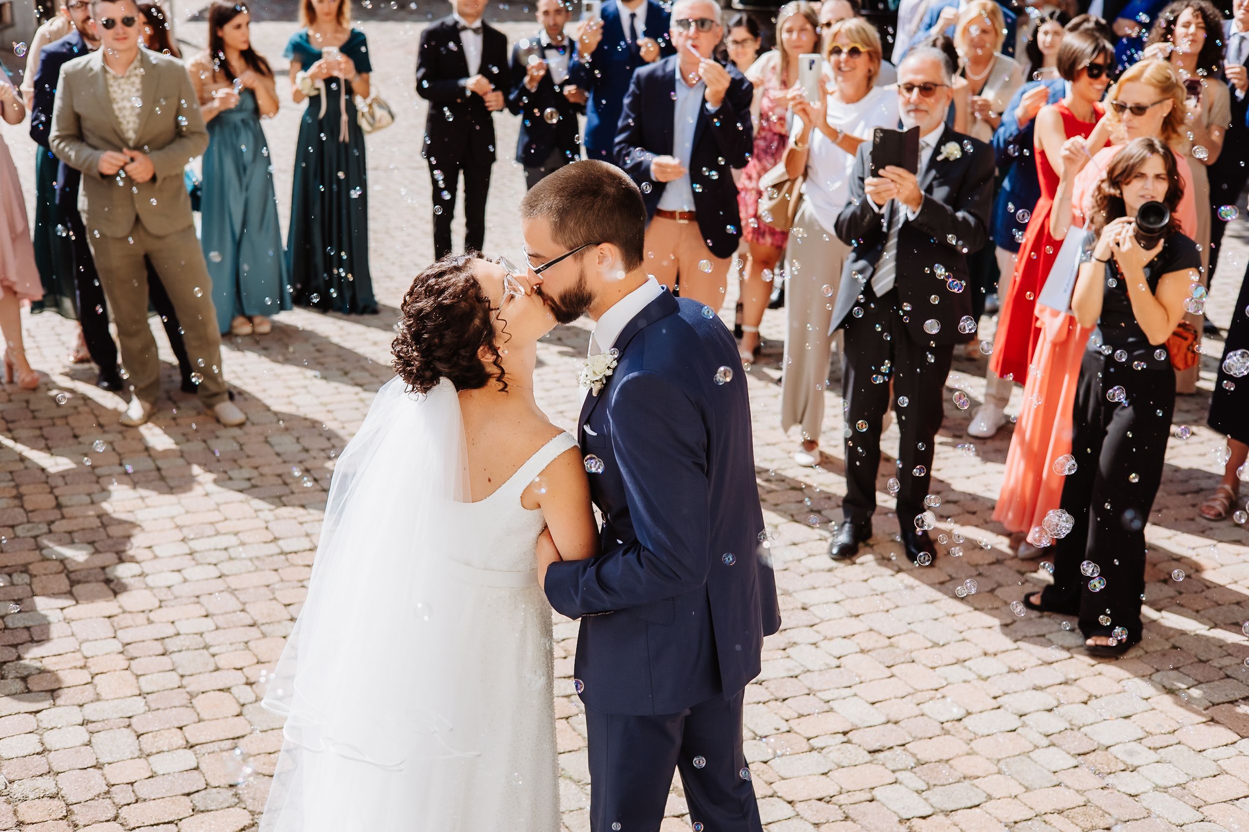 A couple shares their first dance on a cobblestone courtyard surrounded by wedding guests during a sunny celebration.