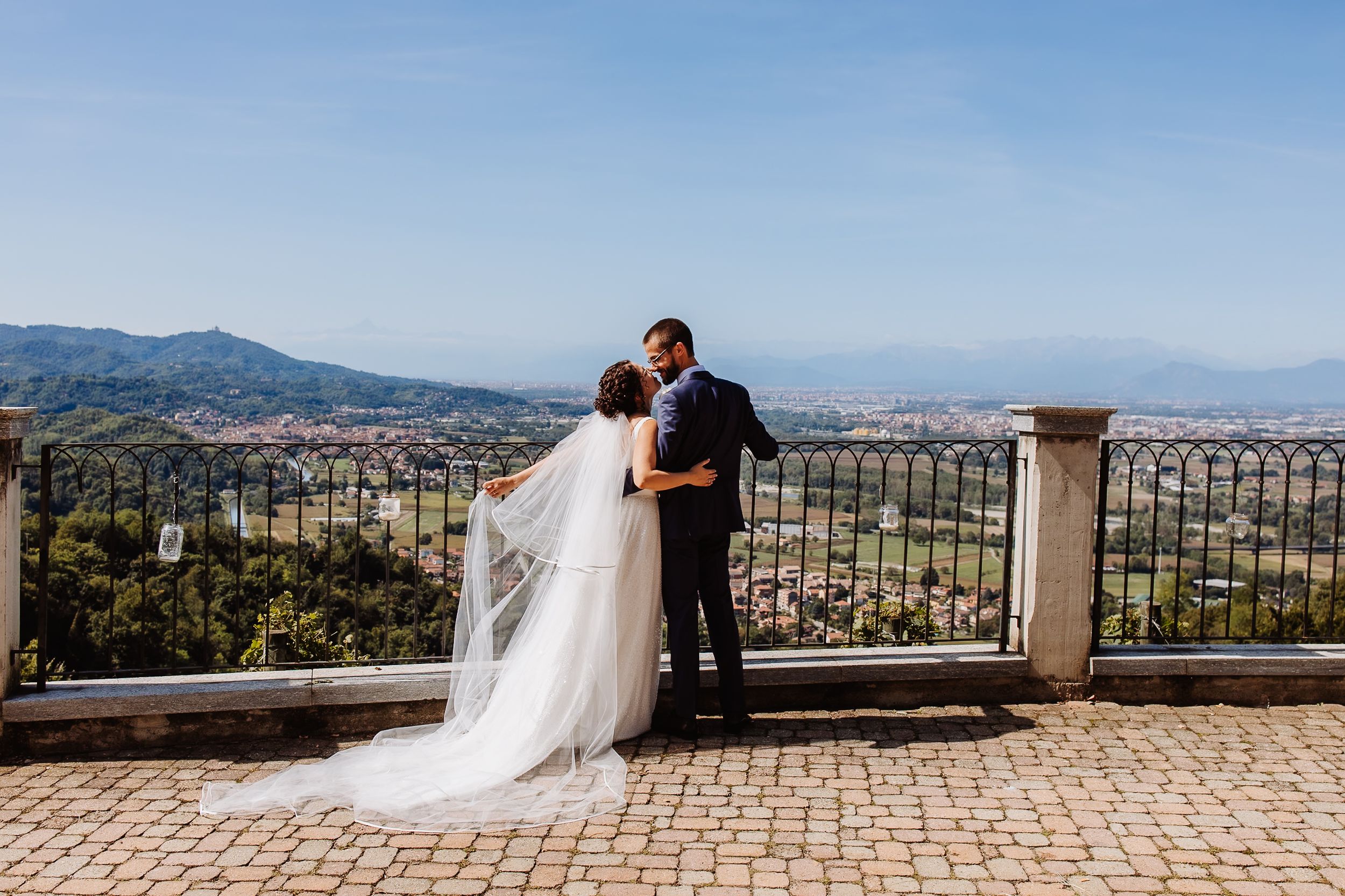 A romantic couple in wedding attire shares a tender moment overlooking a scenic mountain landscape on a sunny day.