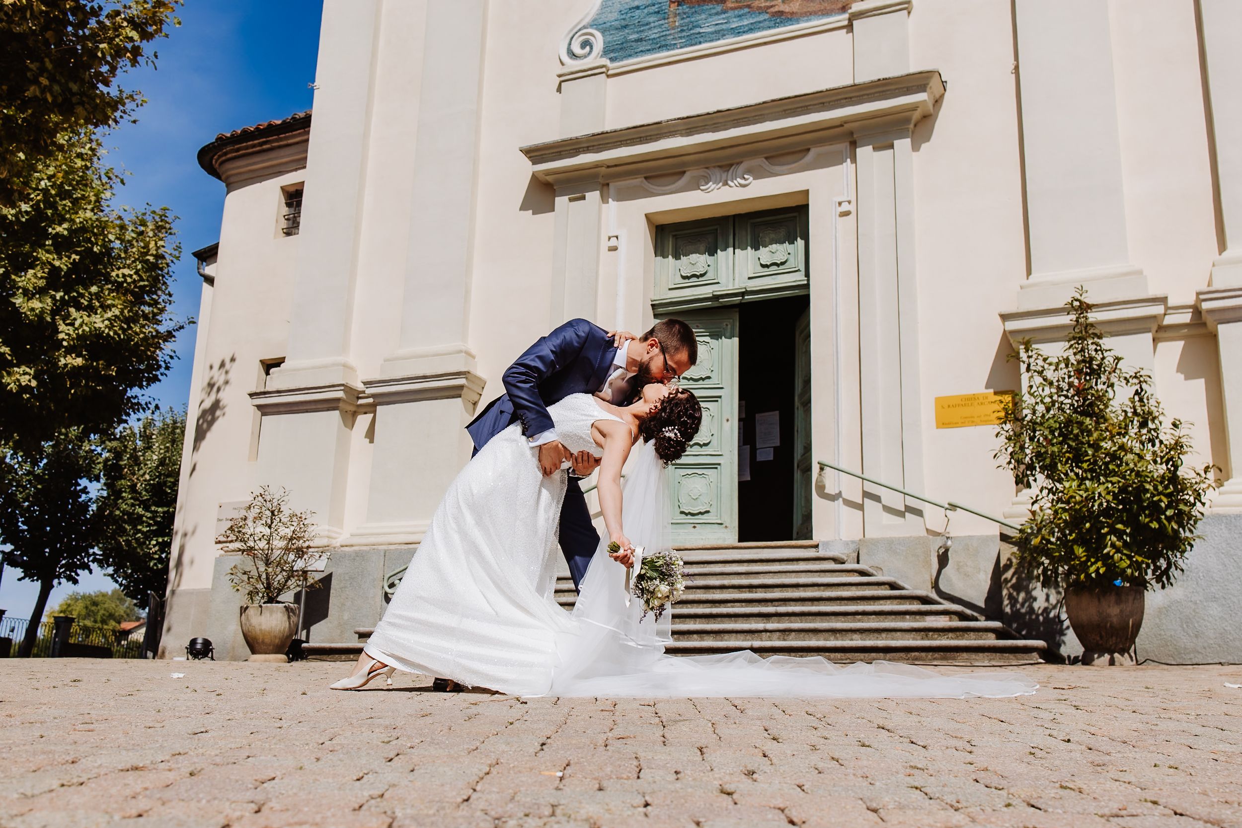 A romantic couple embraces and kisses on white stone steps of an elegant building with classical architecture.