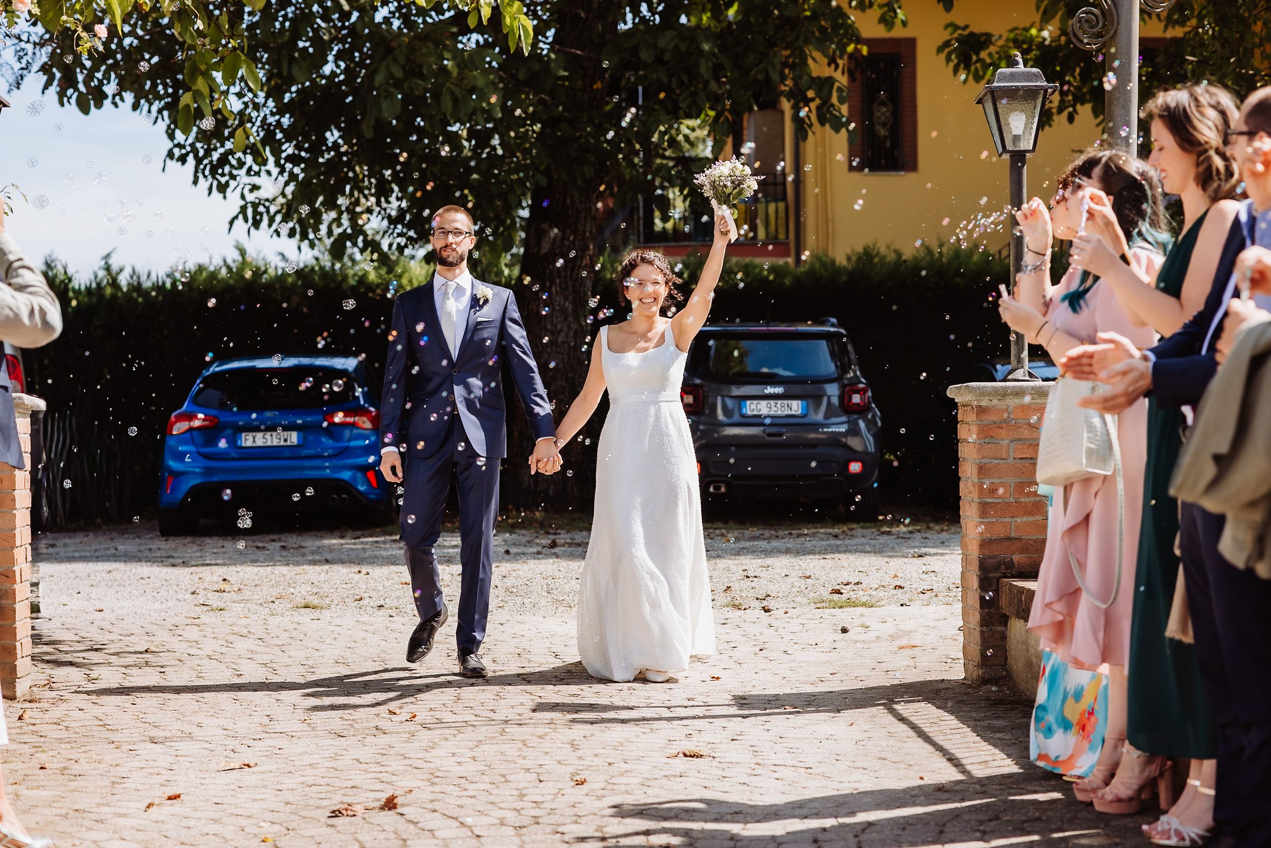 Wedding guests throw confetti and celebrate at an outdoor ceremony in front of a rustic yellow building.