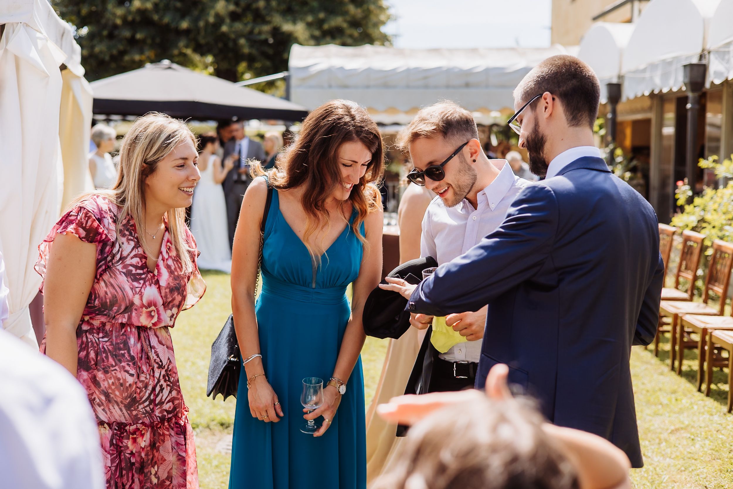 A group of wedding guests dance and socialize outdoors at a sunny reception venue with umbrellas and tables.