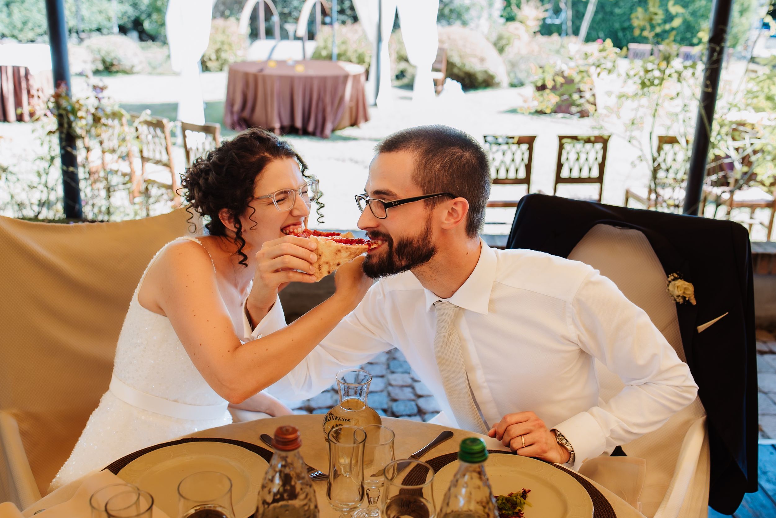 A couple shares a playful moment while feeding each other cake at their outdoor wedding reception.