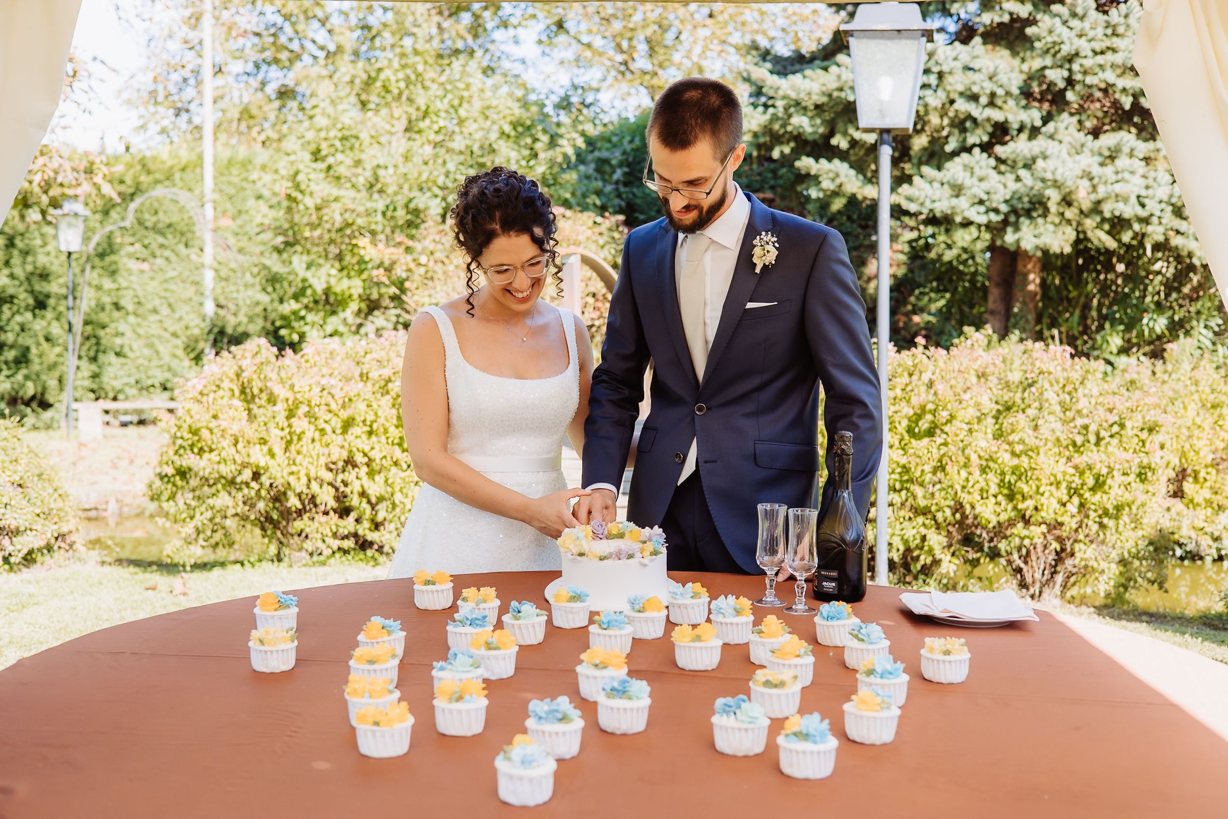 Couple at outdoor wedding reception examining decorated mason jar favors displayed on wooden table.