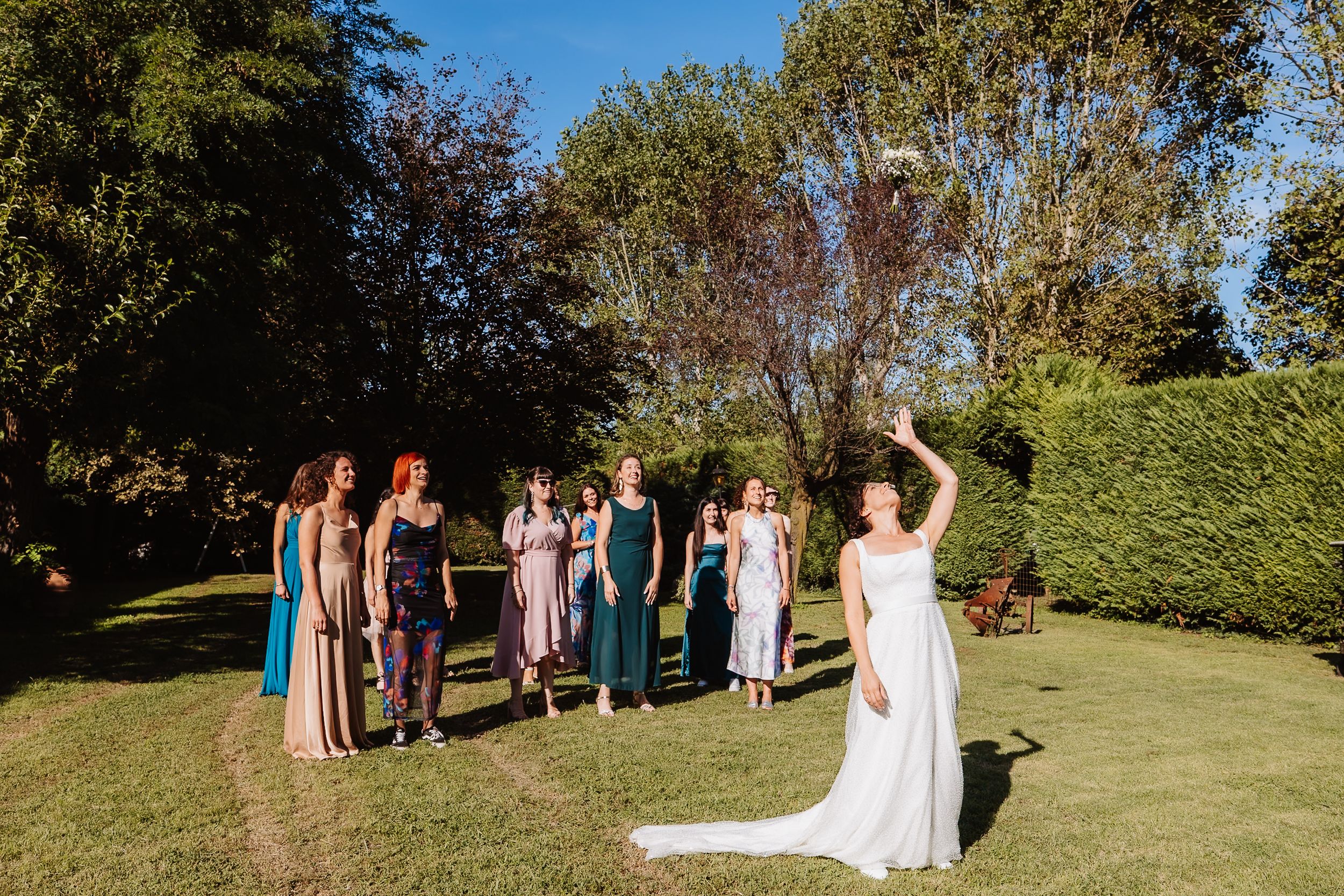 Wedding party members celebrate outdoors on a sunny day in a garden setting with colorful dresses.