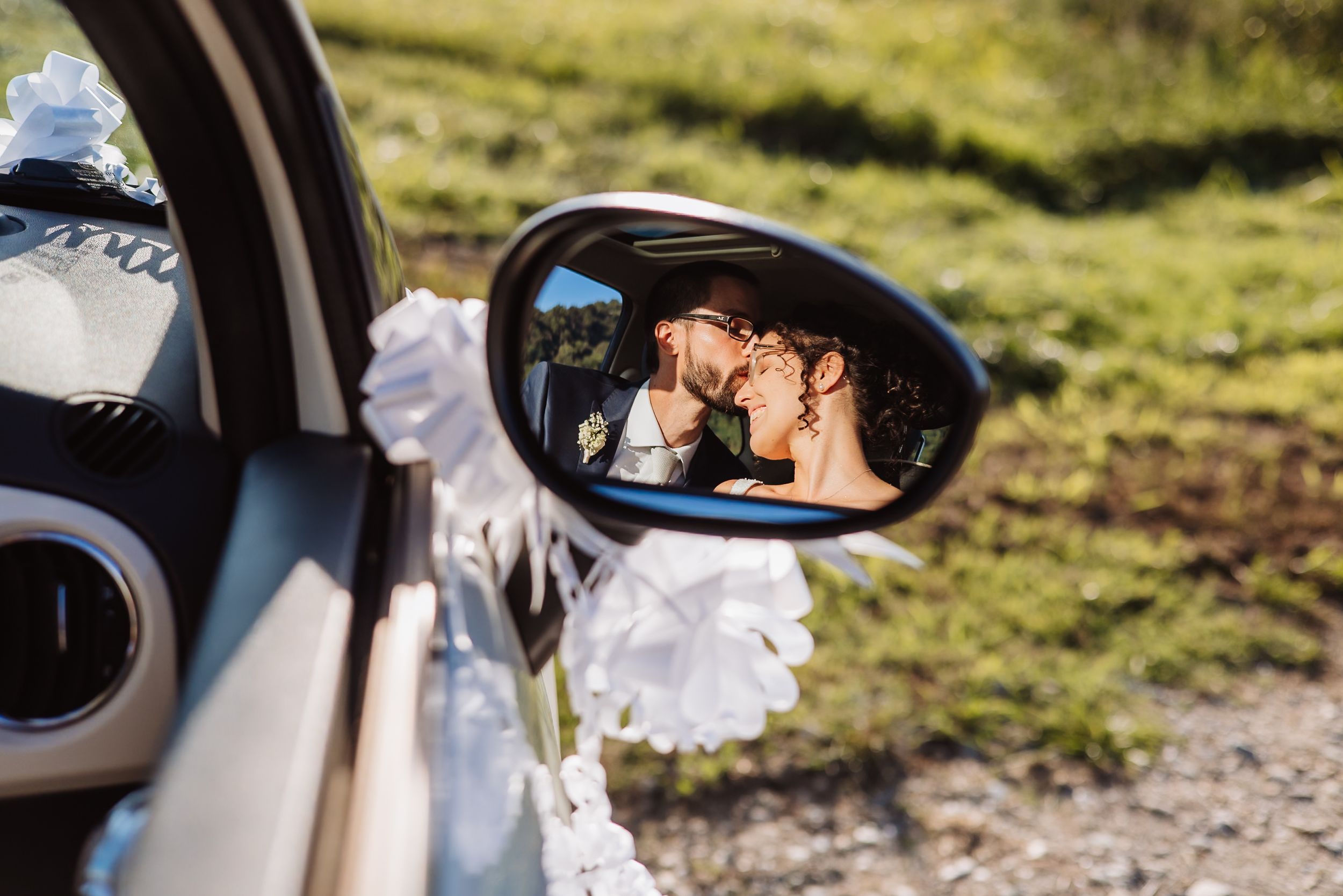 Newlywed couple reflected in car side mirror during outdoor wedding photo session.