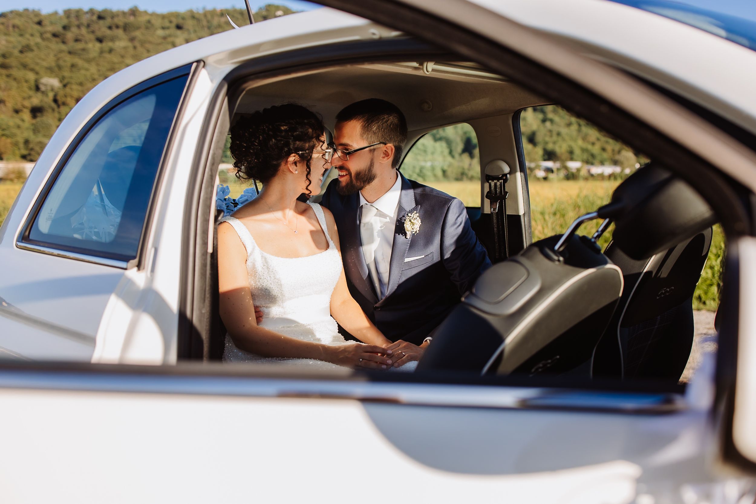 Two people share an intimate moment inside a car on their wedding day, dressed in formal white attire.