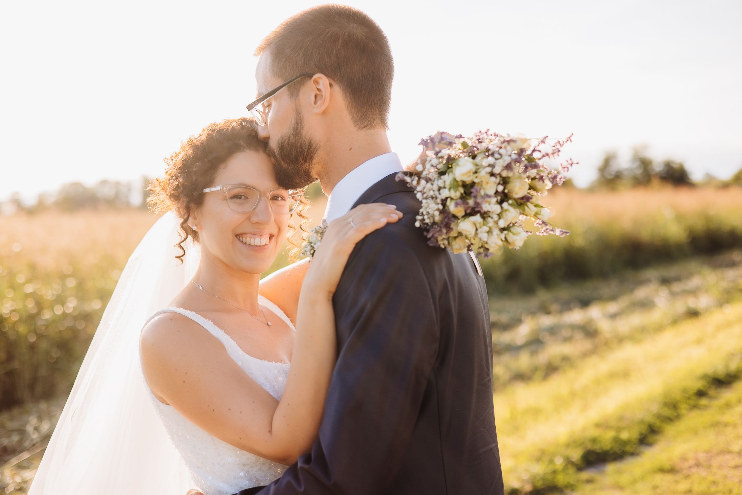 A romantic couple in wedding attire embrace during golden hour in a scenic outdoor field setting.