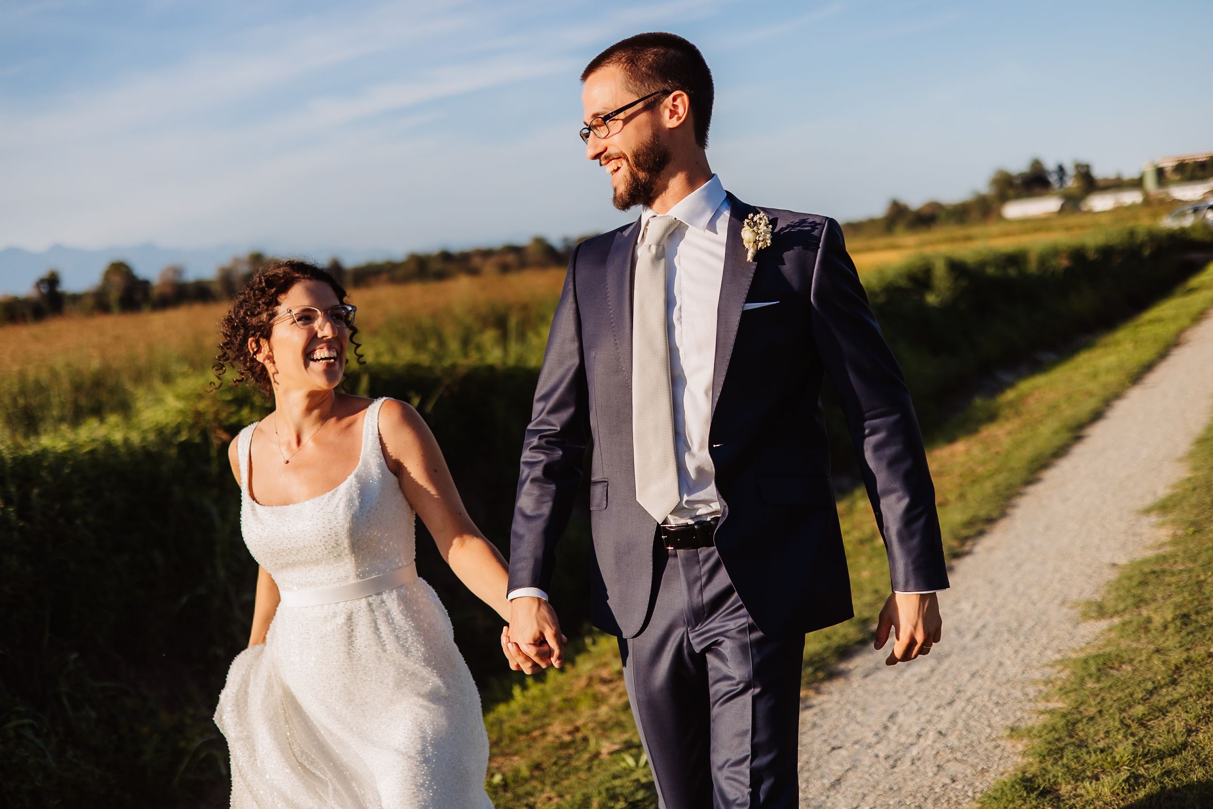 A couple in wedding attire strolls hand in hand down a rural path at golden hour.