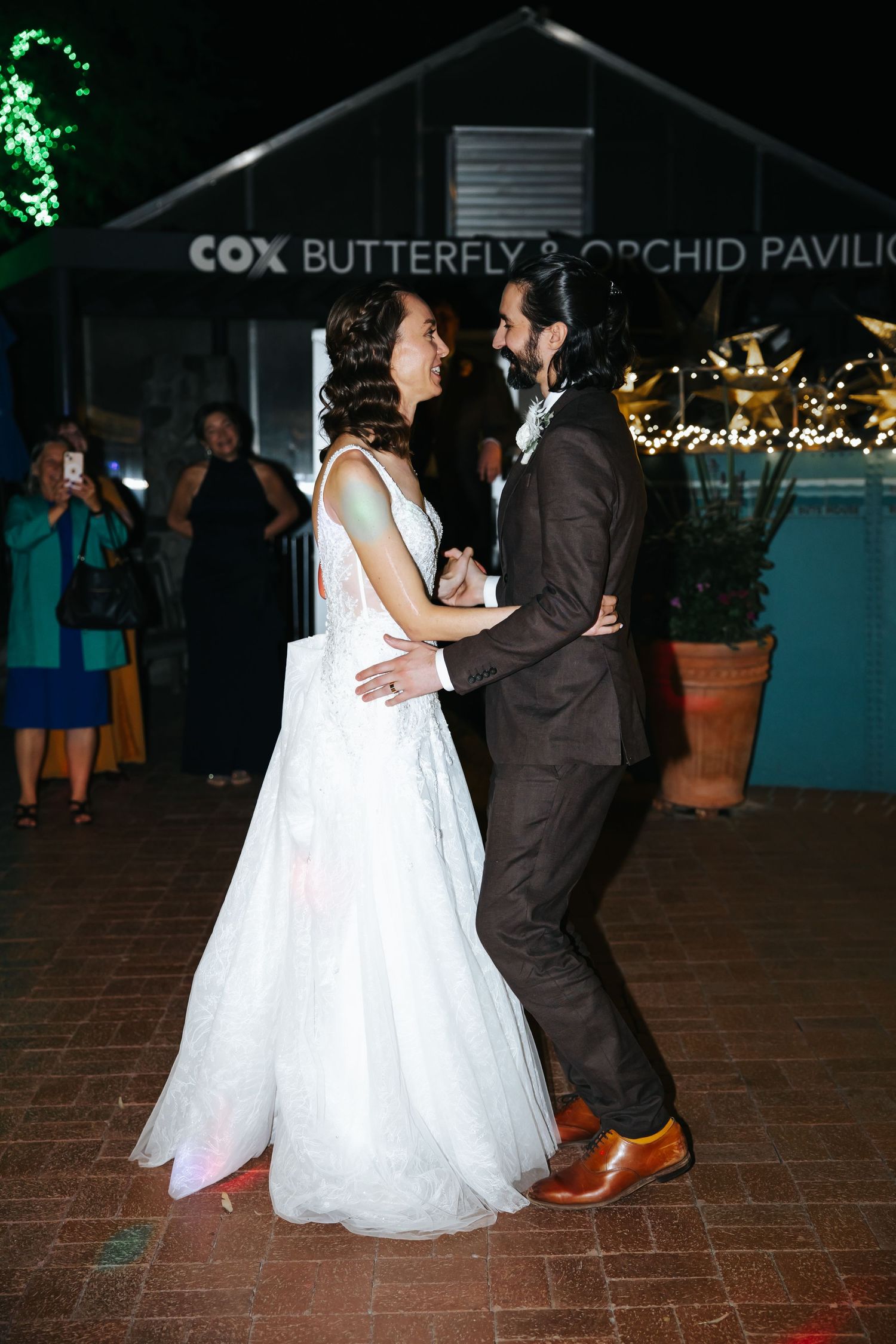 A couple dances together at an evening wedding reception on a brick patio with twinkling lights in the background.