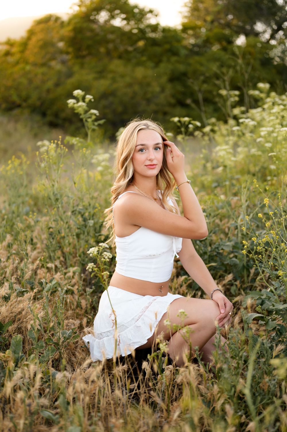 Blonde high school senior wearing white dress sits in wildflowers during outdoor senior portrait session Ventura photographer