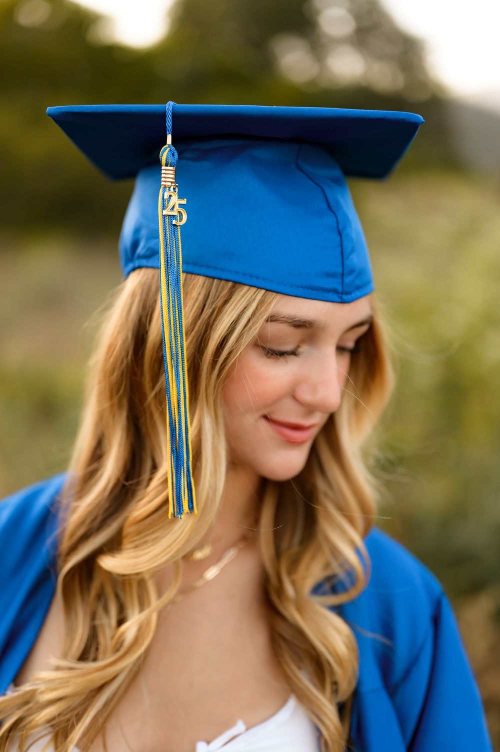 Graduating high school senior wearing white dress and cap and gown poses during outdoor senior portrait session Ventura photographer