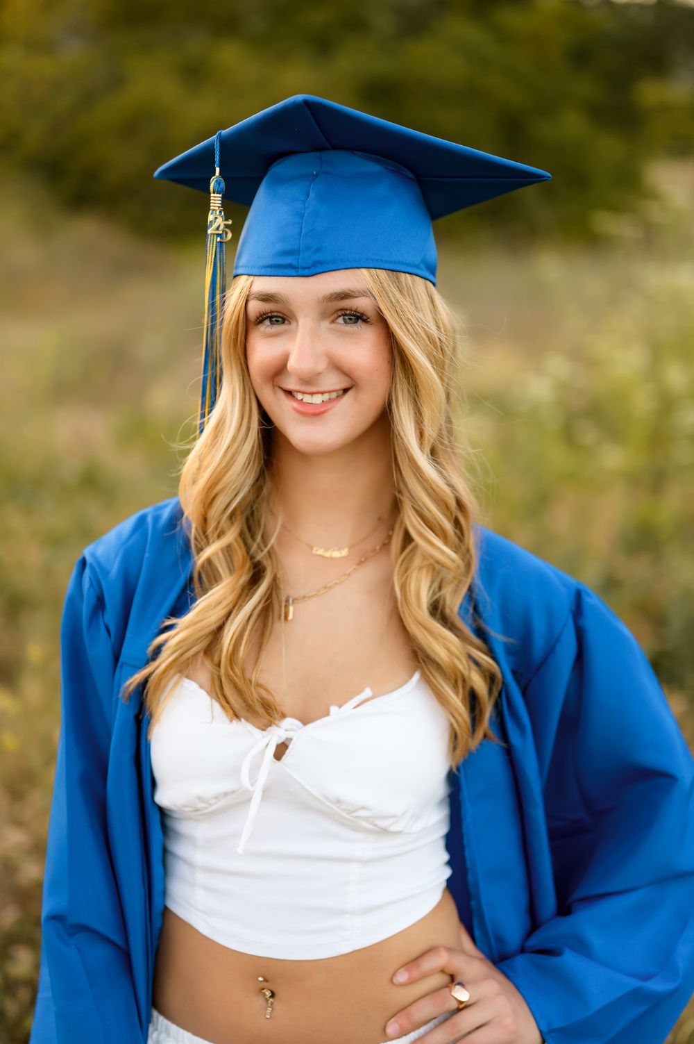 Graduating high school senior wearing white dress and cap and gown poses during outdoor senior portrait session Ventura photographer