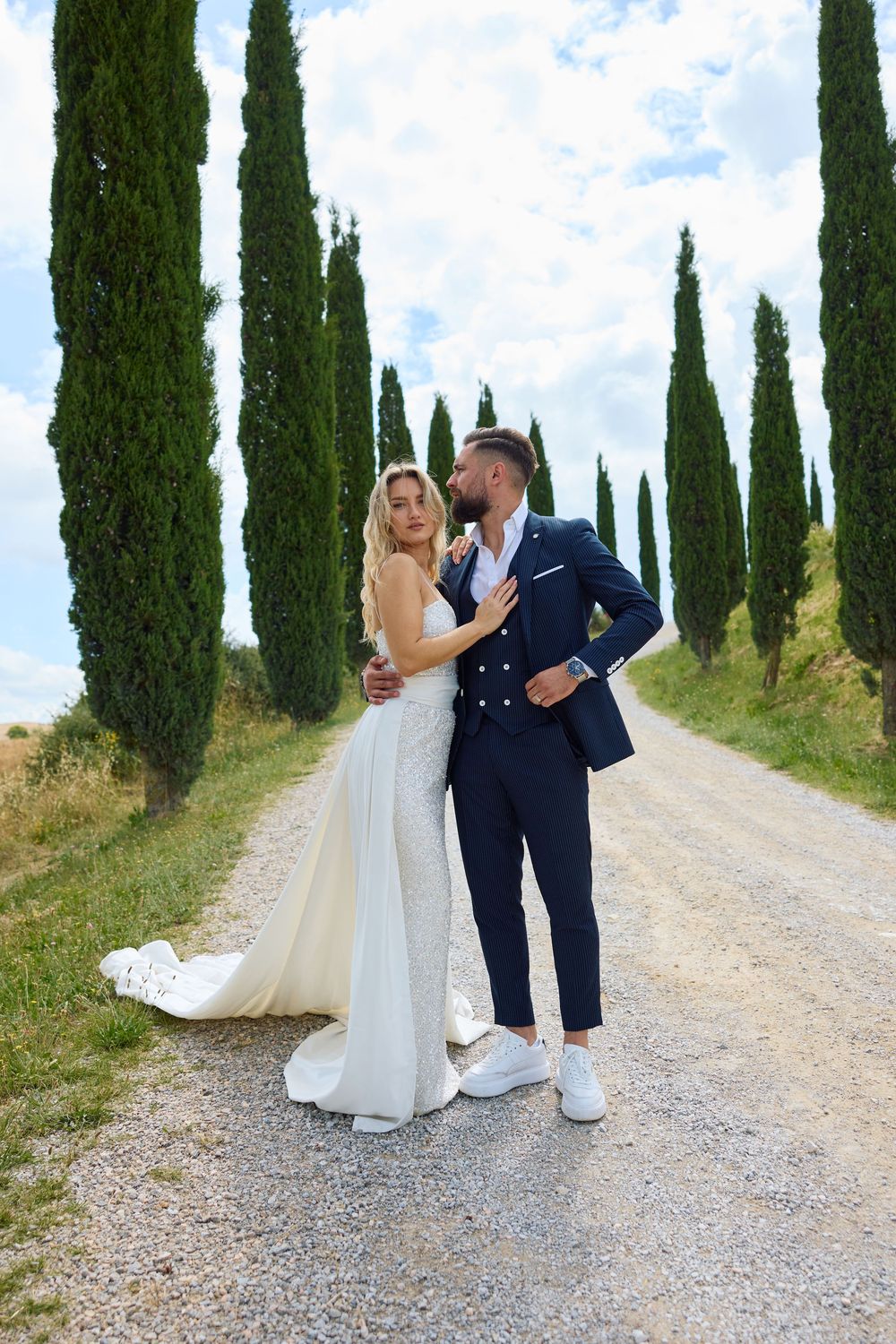 A romantic wedding couple poses along a cypress tree-lined gravel path in the Tuscan countryside.
